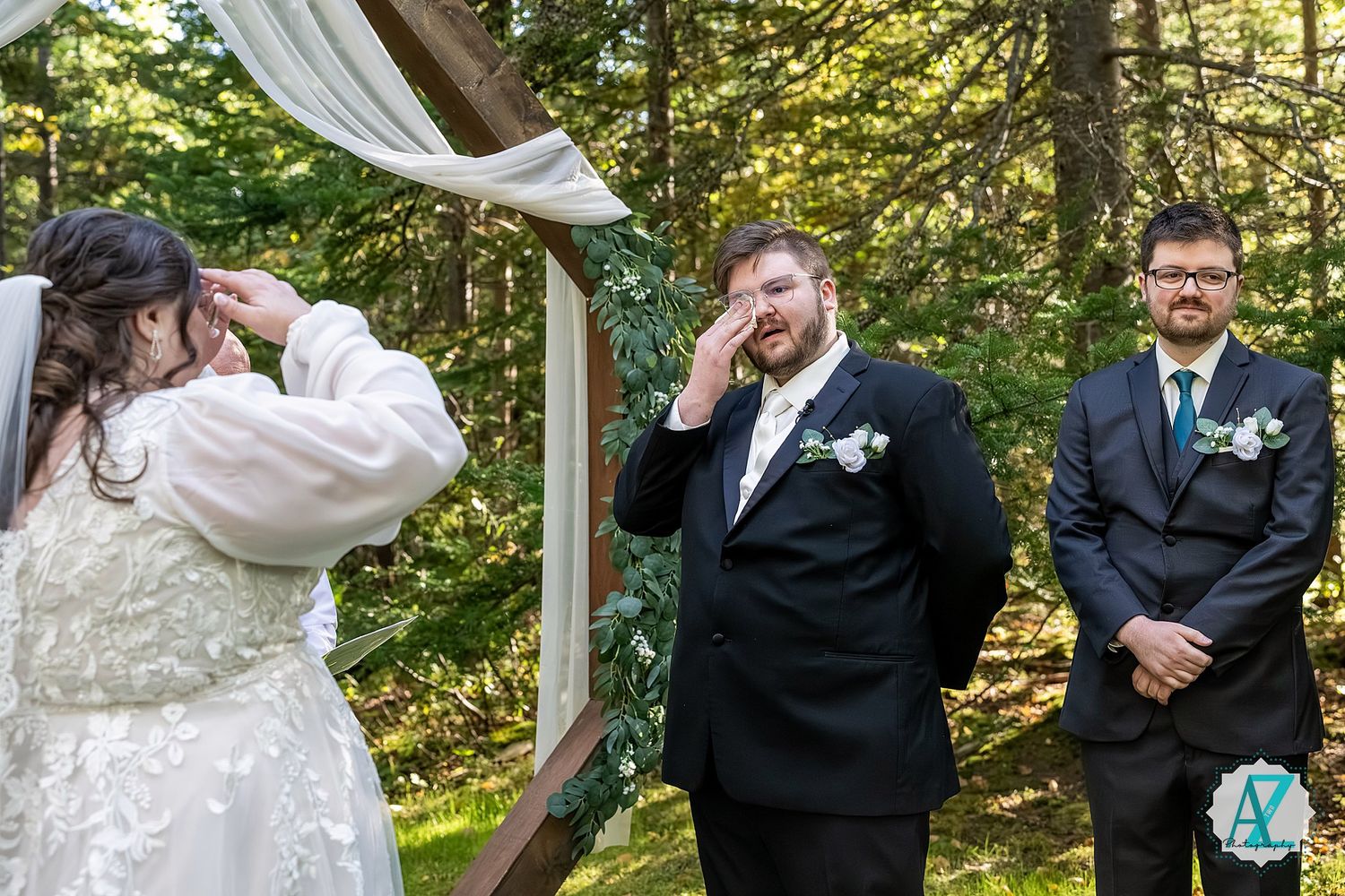 Wedding party poses joyfully in a sun-dappled forest setting with flowing white fabric and formal attire.