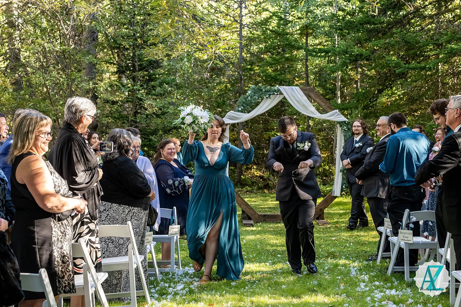 Wedding guests gather outdoors in a wooded setting with white chairs arranged for a ceremony.