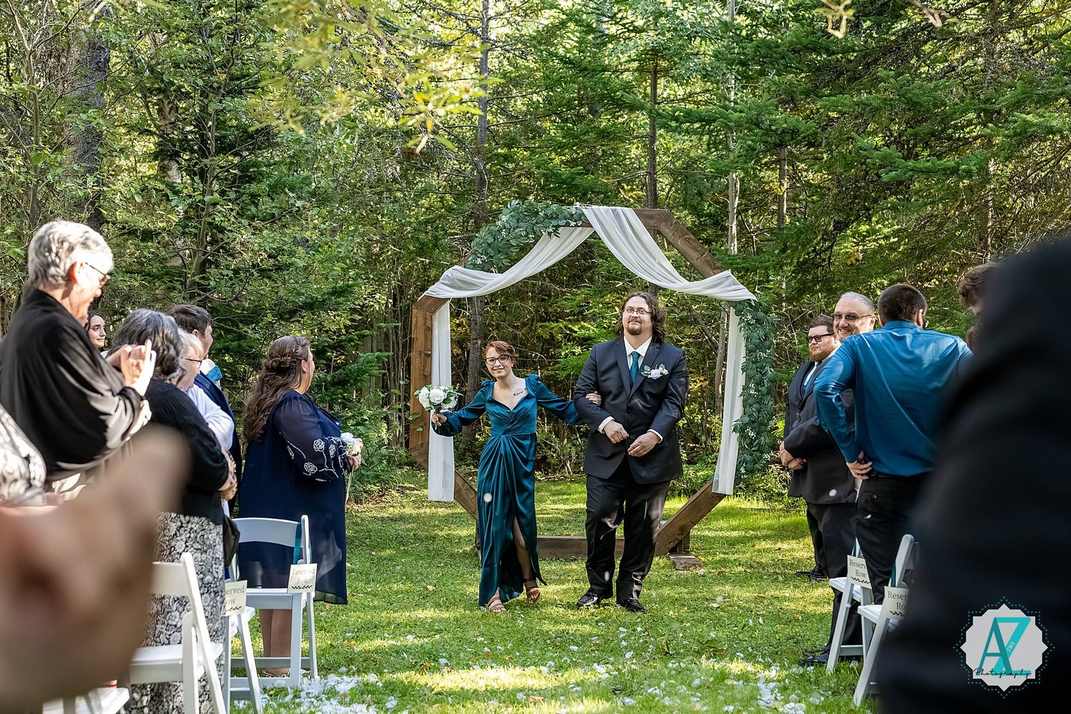 Outdoor wedding ceremony in a wooded setting with guests seated on wooden benches and a white arch at the front.