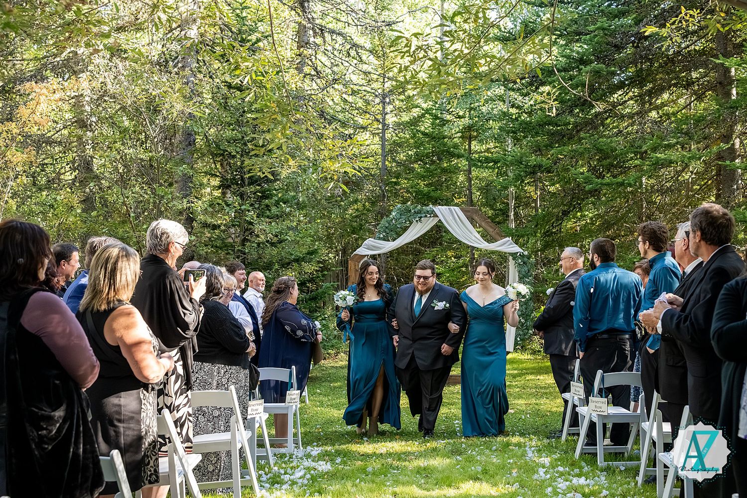 A small outdoor wedding ceremony takes place in a lush forest setting with white chairs and a decorative arch.