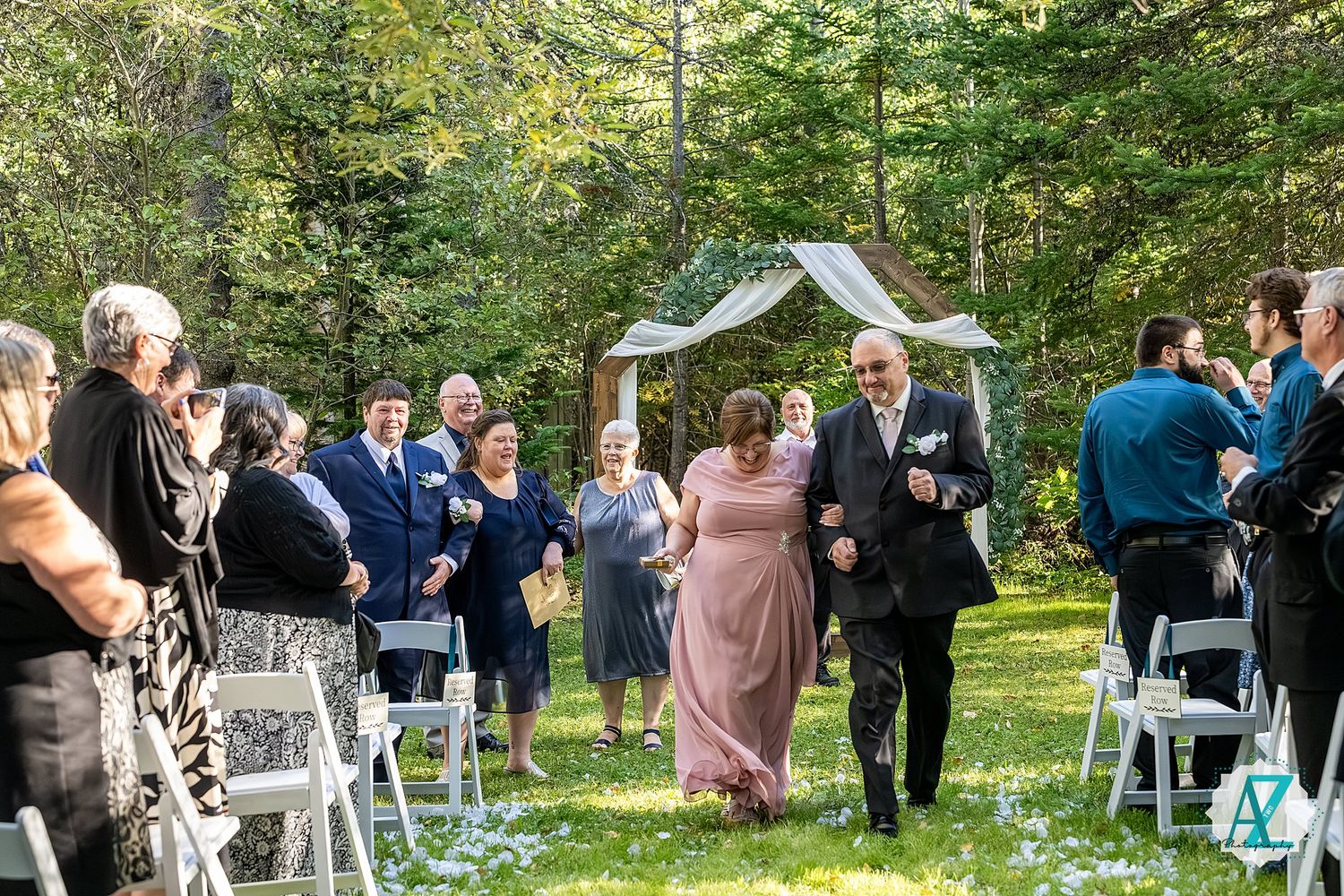 A wedding ceremony takes place in a lush garden setting with white chairs and a wooden arch surrounded by greenery.