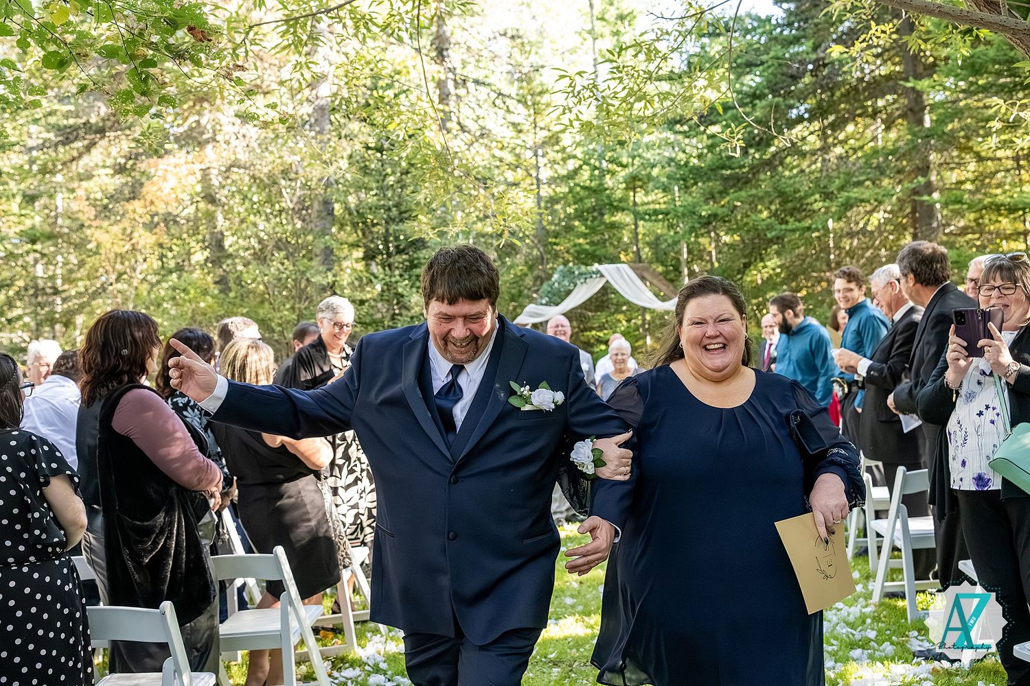 Outdoor wedding celebration in a forest clearing with guests mingling among white chairs.