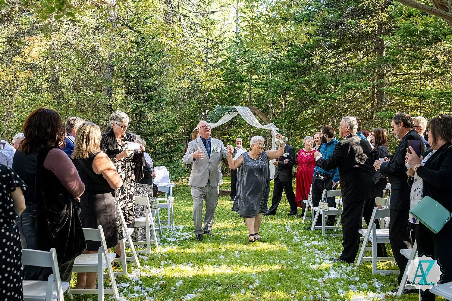 Forest wedding ceremony setup with white chairs and guests gathering among tall trees.