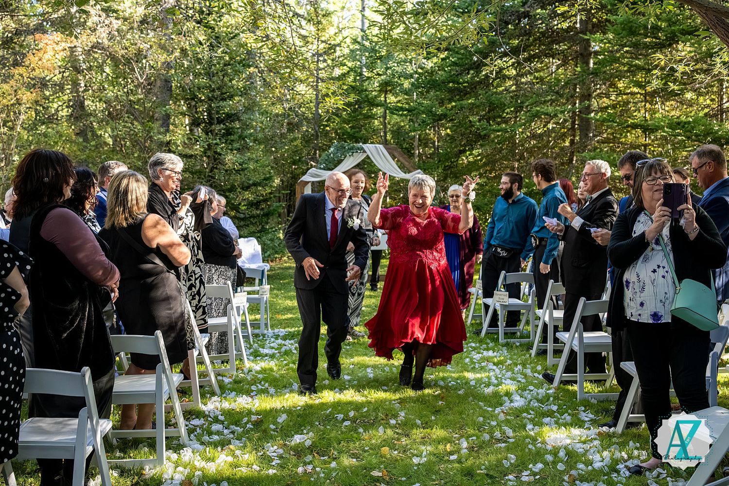 Guests gathered outdoors for a garden wedding ceremony with white chairs arranged on a sunny lawn surrounded by trees.