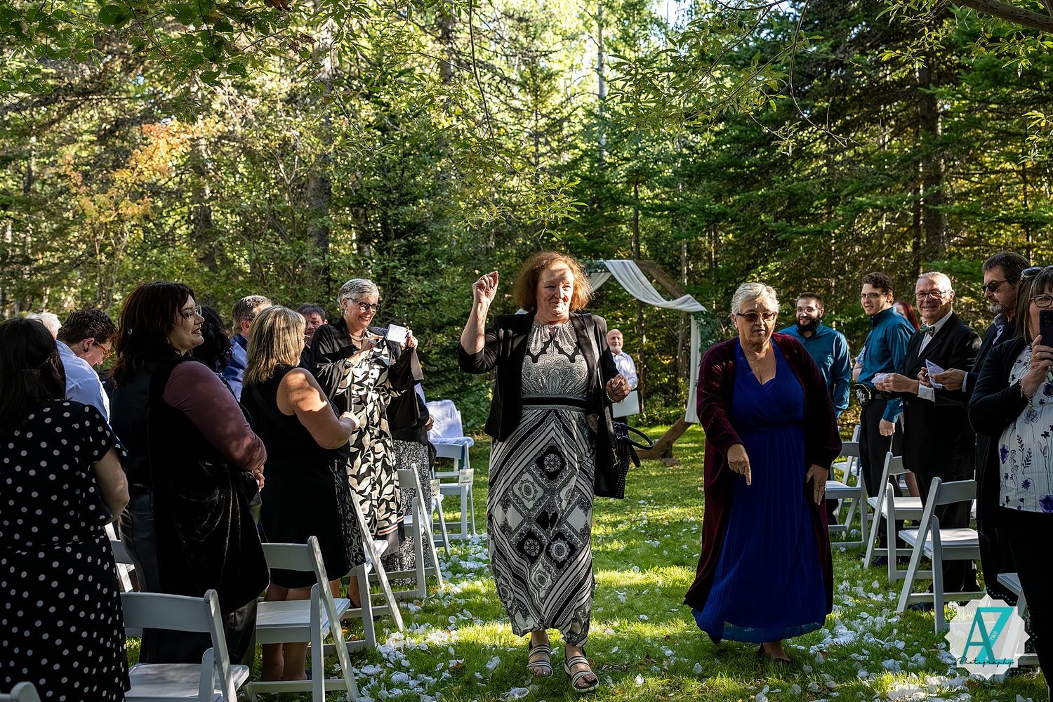 Wedding guests gather outdoors for a ceremony in a lush garden setting with white chairs arranged in rows.