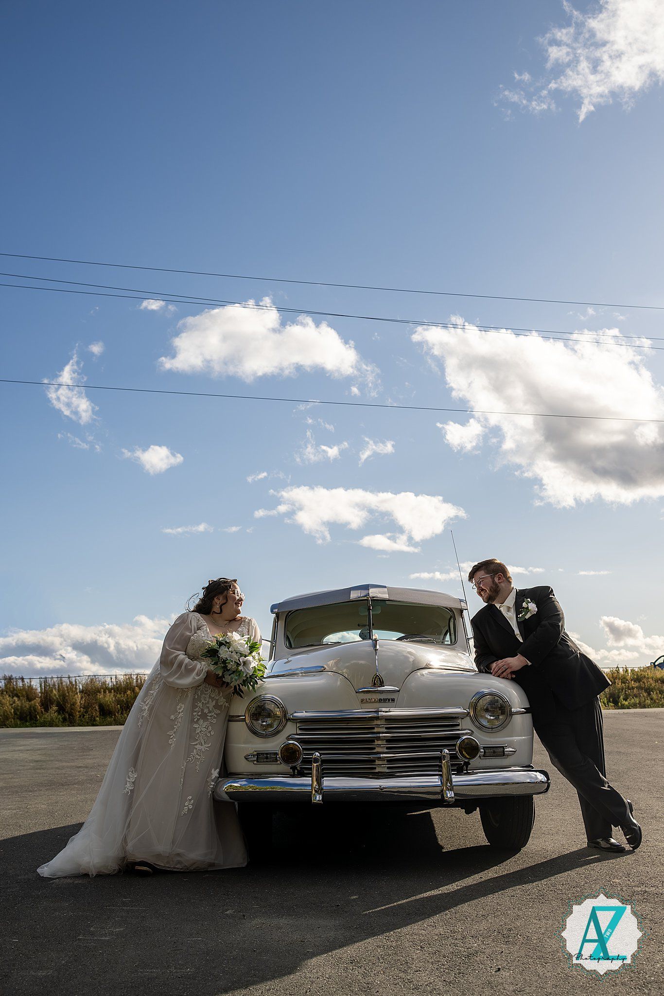 A vintage white Chevrolet car parked on a road with a couple posing playfully in front of it on a sunny day.