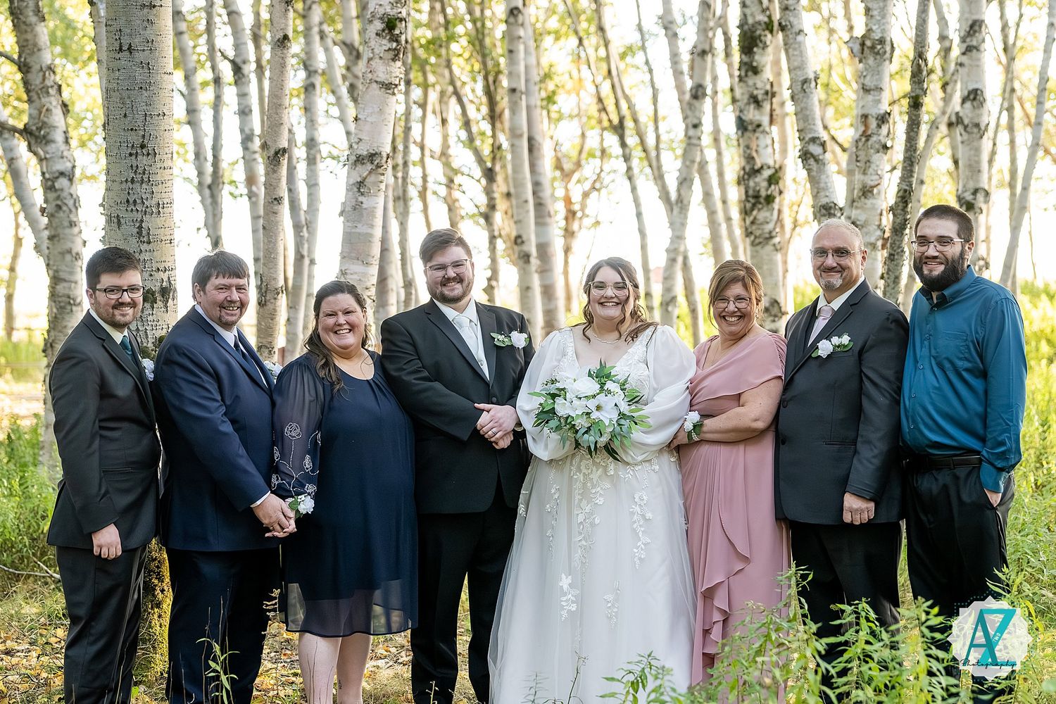 Large wedding party poses together between tall white birch trees in a wooded outdoor setting.