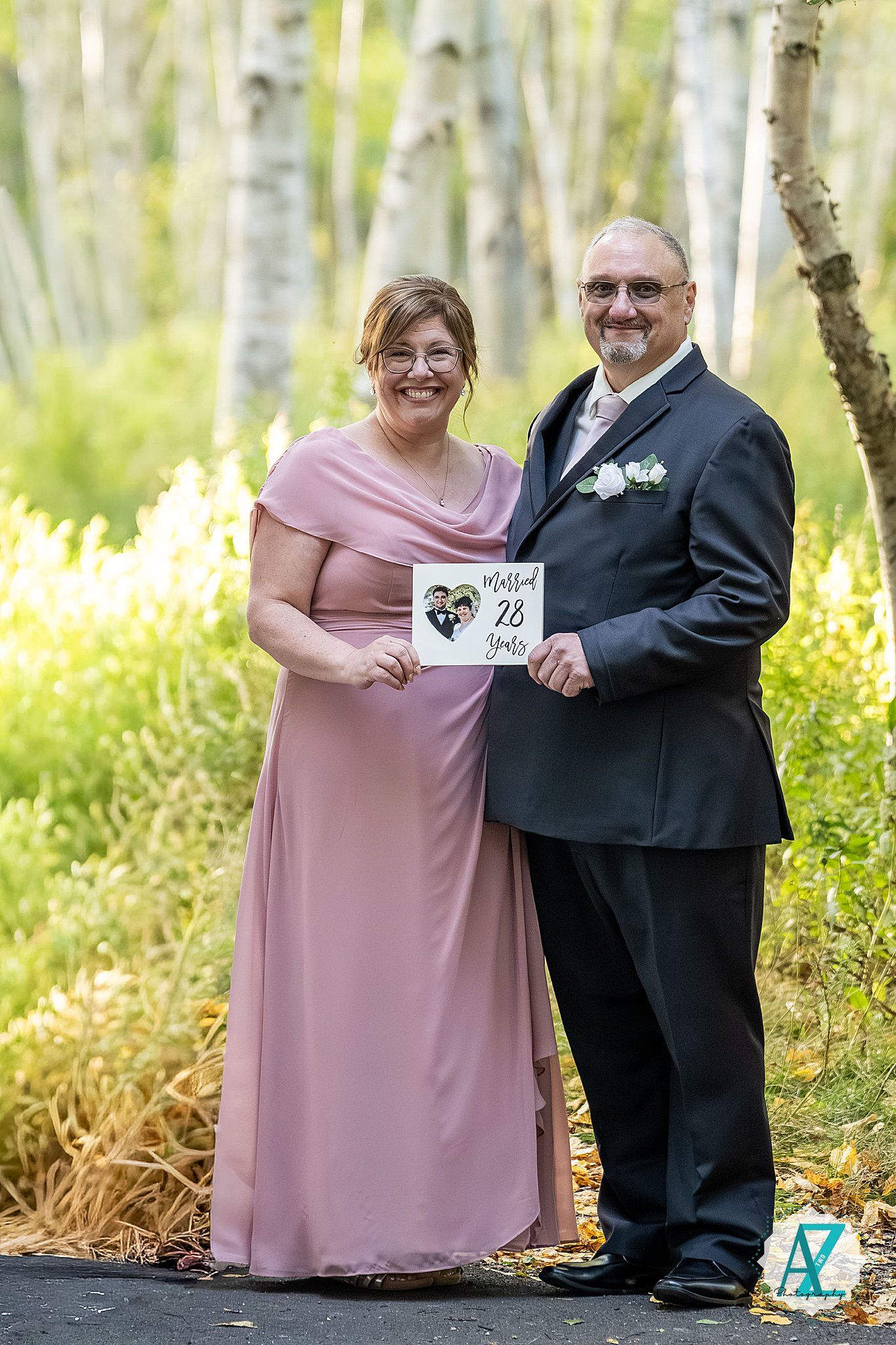 A couple in formal attire poses in a birch forest holding a wedding sign together.