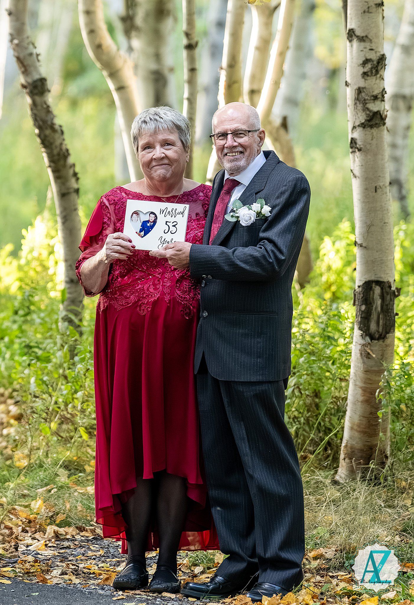 A couple in formal wear poses among birch trees holding a heart-shaped sign.