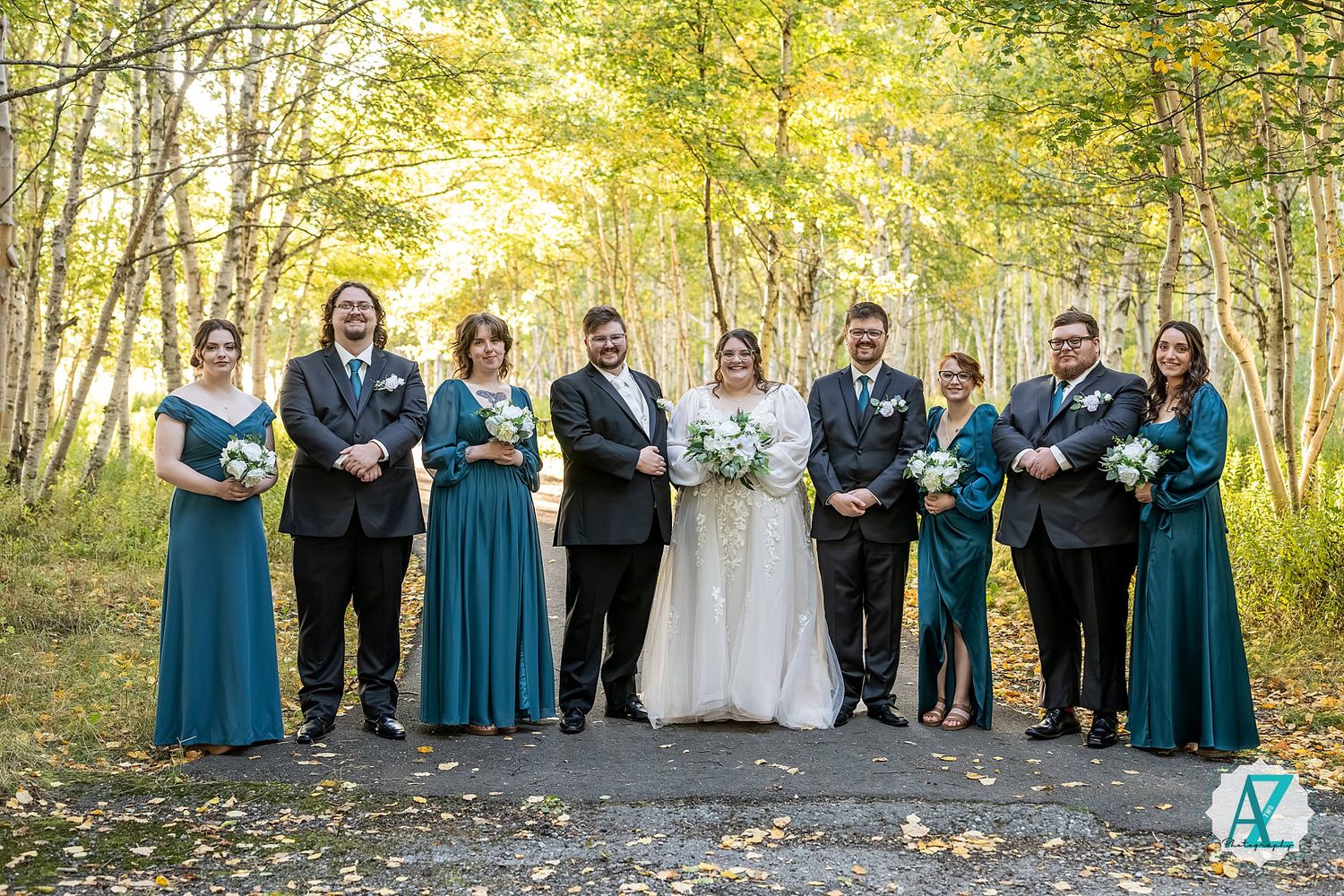 Wedding party in formal attire poses together on wooded path during autumn outdoor celebration.