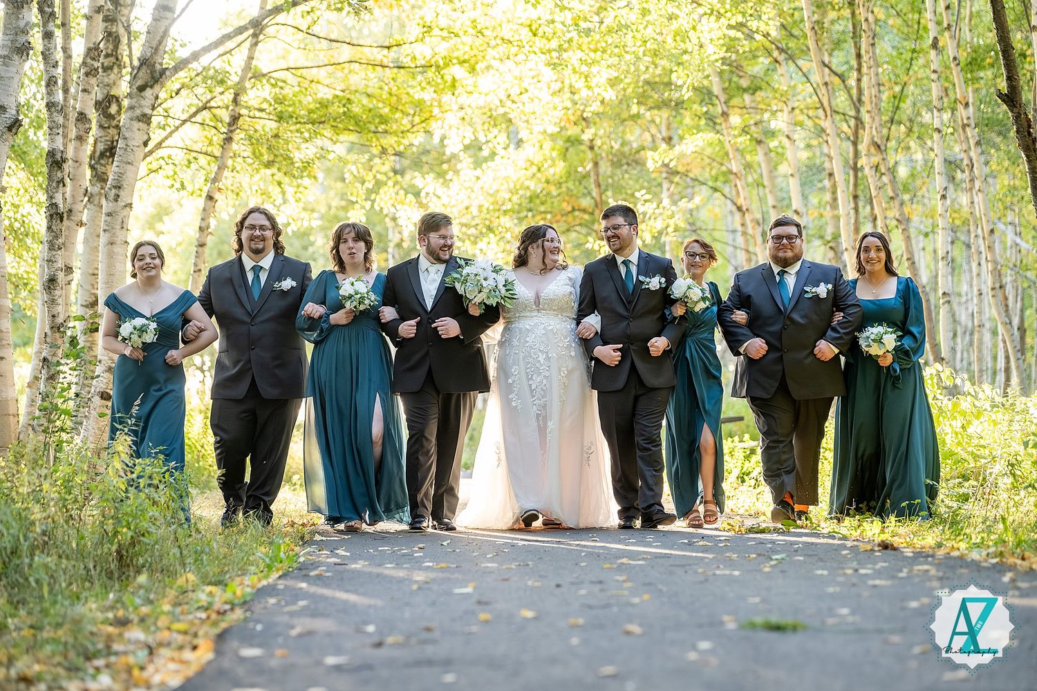 Wedding party poses together on a tree-lined path during autumn with bridesmaids in teal dresses and groomsmen in dark suits.