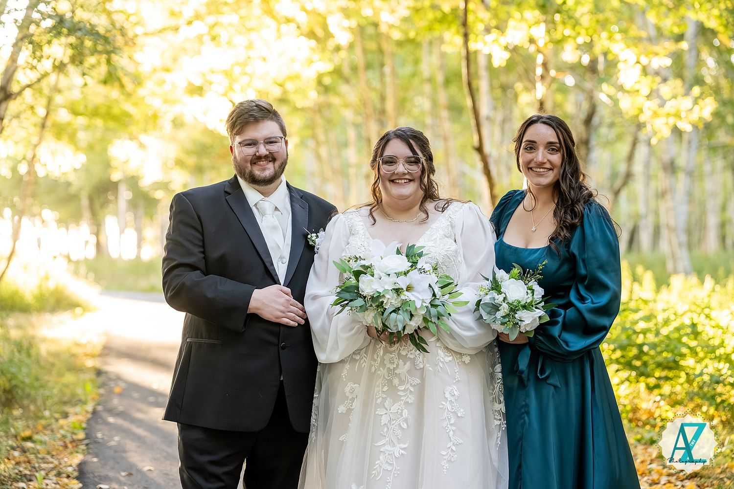 Wedding party members in teal and formal attire pose together in a sunlit autumn forest setting.