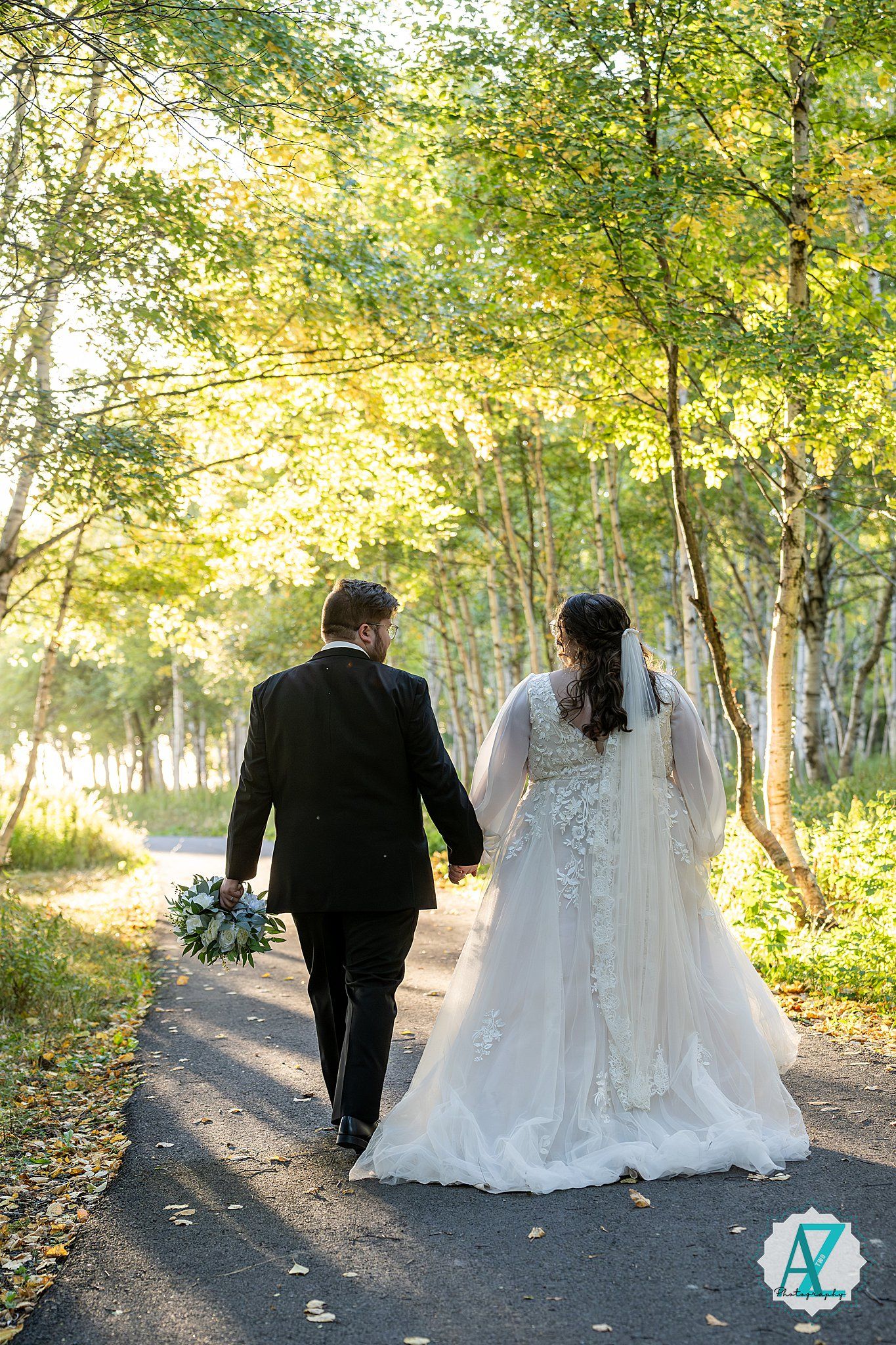 Newlyweds walk hand in hand down a sunlit forest path surrounded by birch trees.