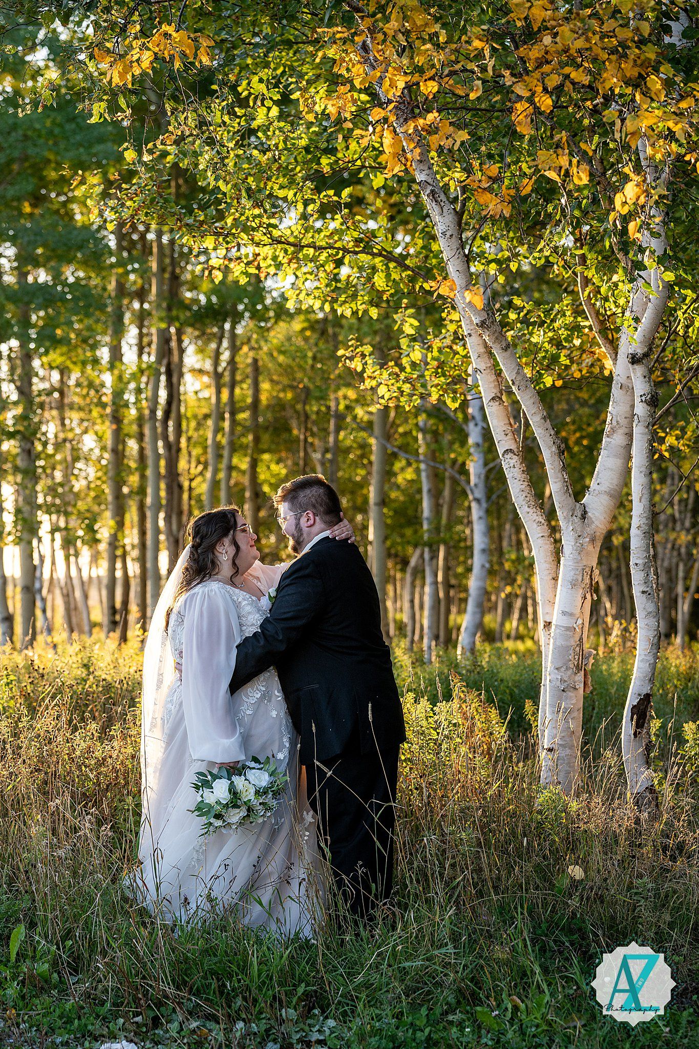 Wedding couple stands among white birch trees in a golden autumn forest setting during sunset.