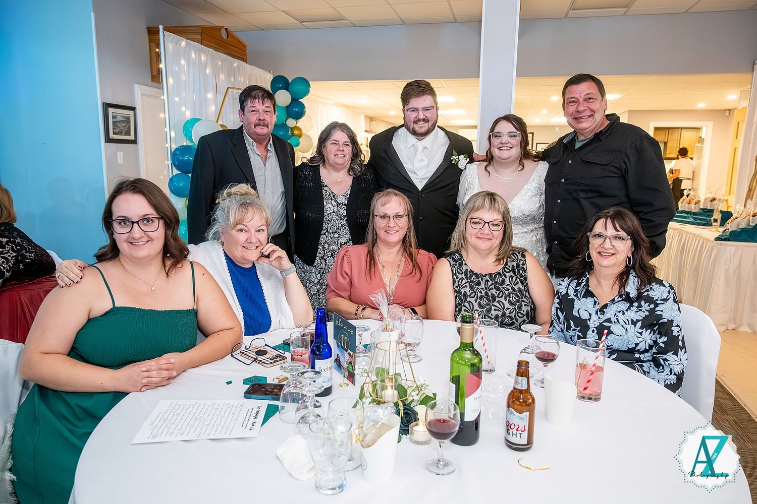 A group of guests in formal attire gathered around a white table at a celebratory event with drinks and decorations.