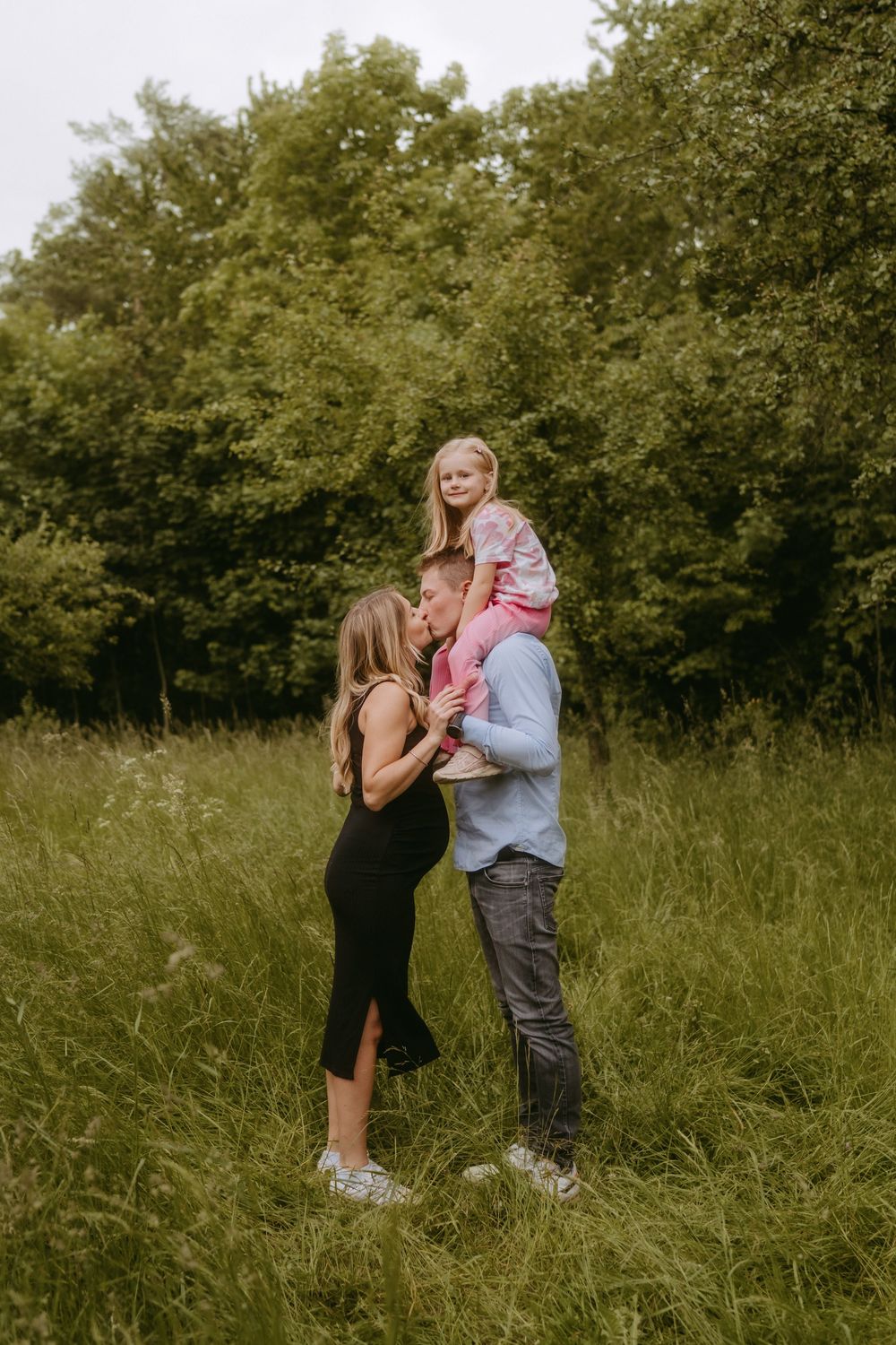 A couple shares a tender moment in a grassy field surrounded by trees.