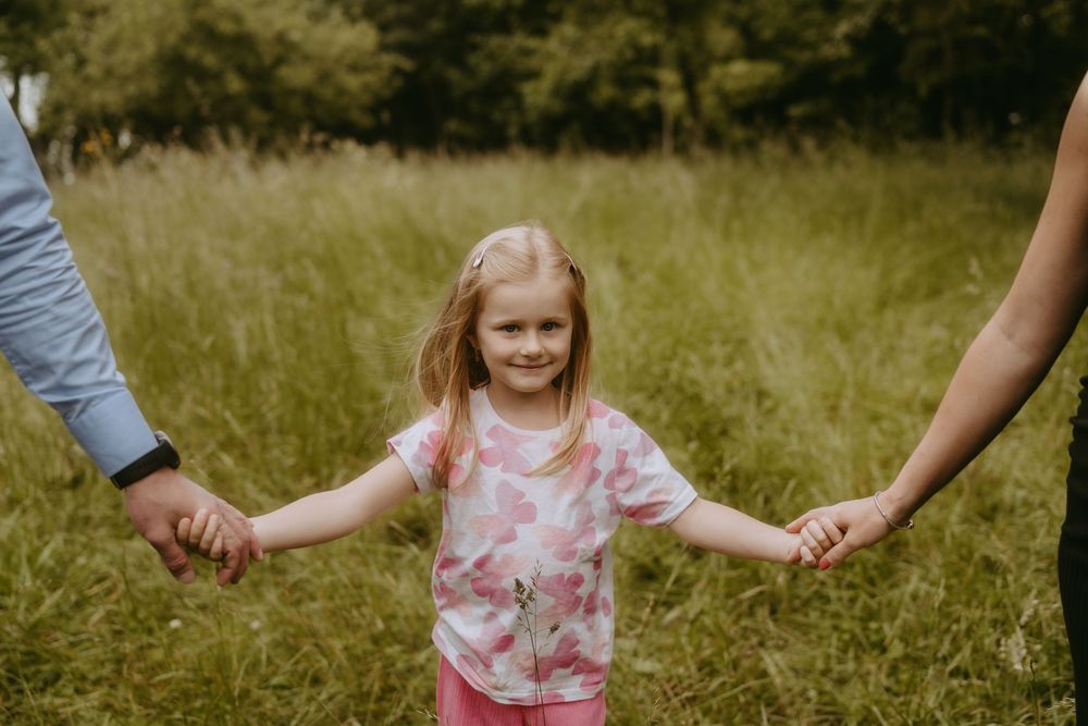 Child in pink floral dress holding hands with adults while walking through tall grass.