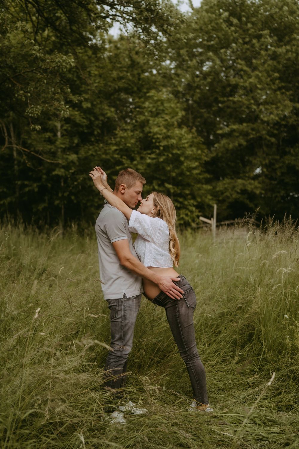 Couple shares tender moments in a sunlit field with tall grass and trees in the background.