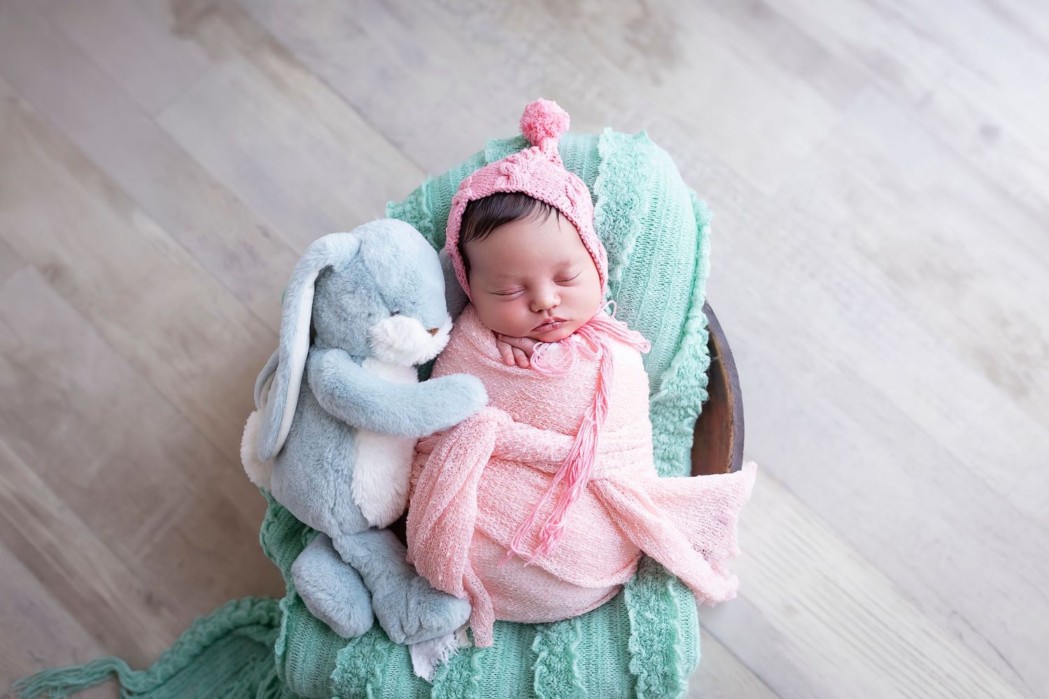 baby girl lays wrapped on a blanket on a wooden bowl shes wearing a pompom bonnet and has a stuffed animal bunny