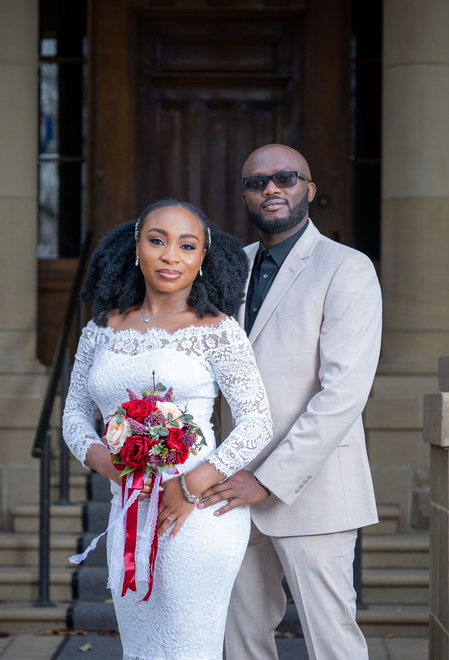 Wedding couple in white lace dress and cream suit posing with red bouquet on stone building steps.