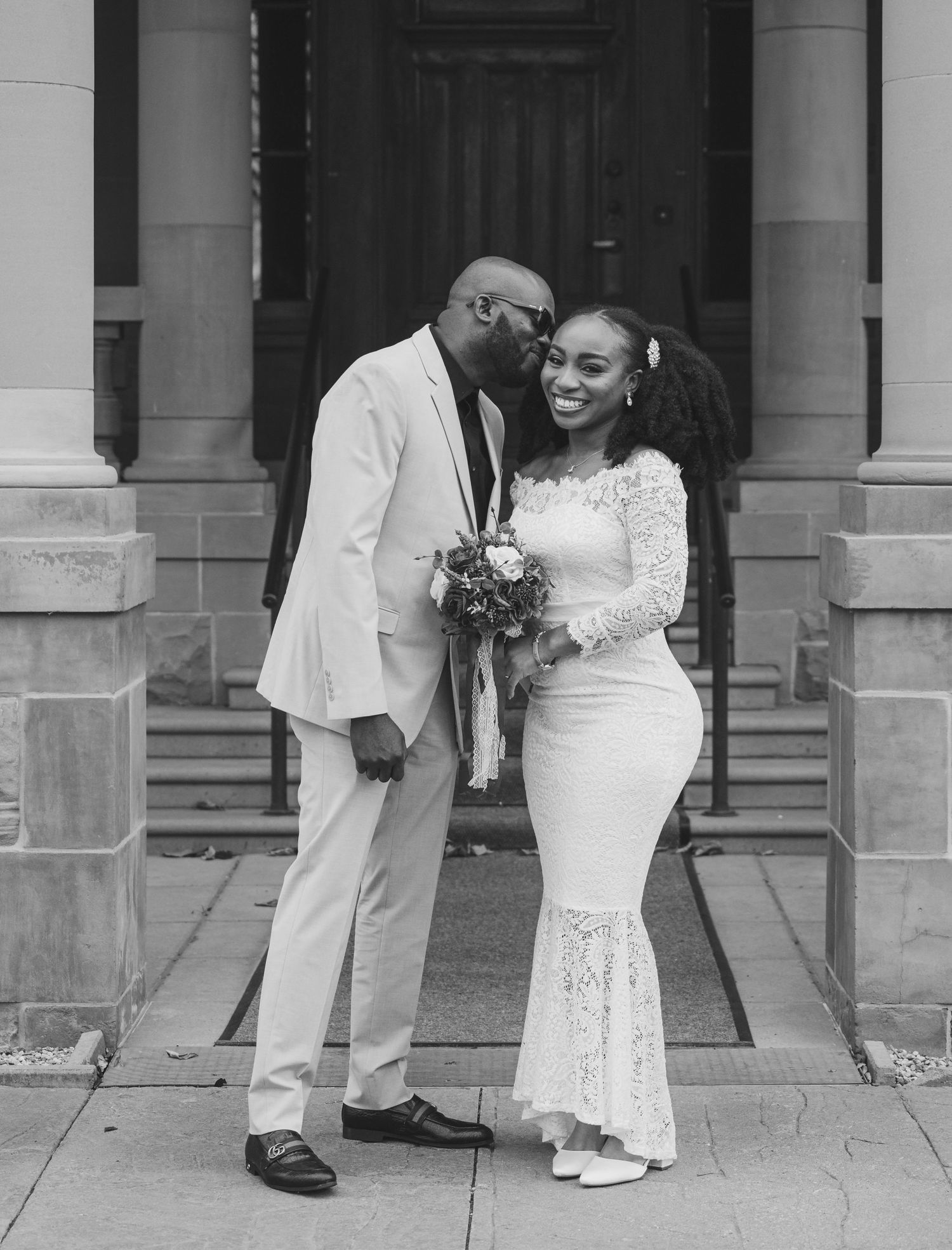 Black and white wedding portrait of couple in elegant attire on building steps.