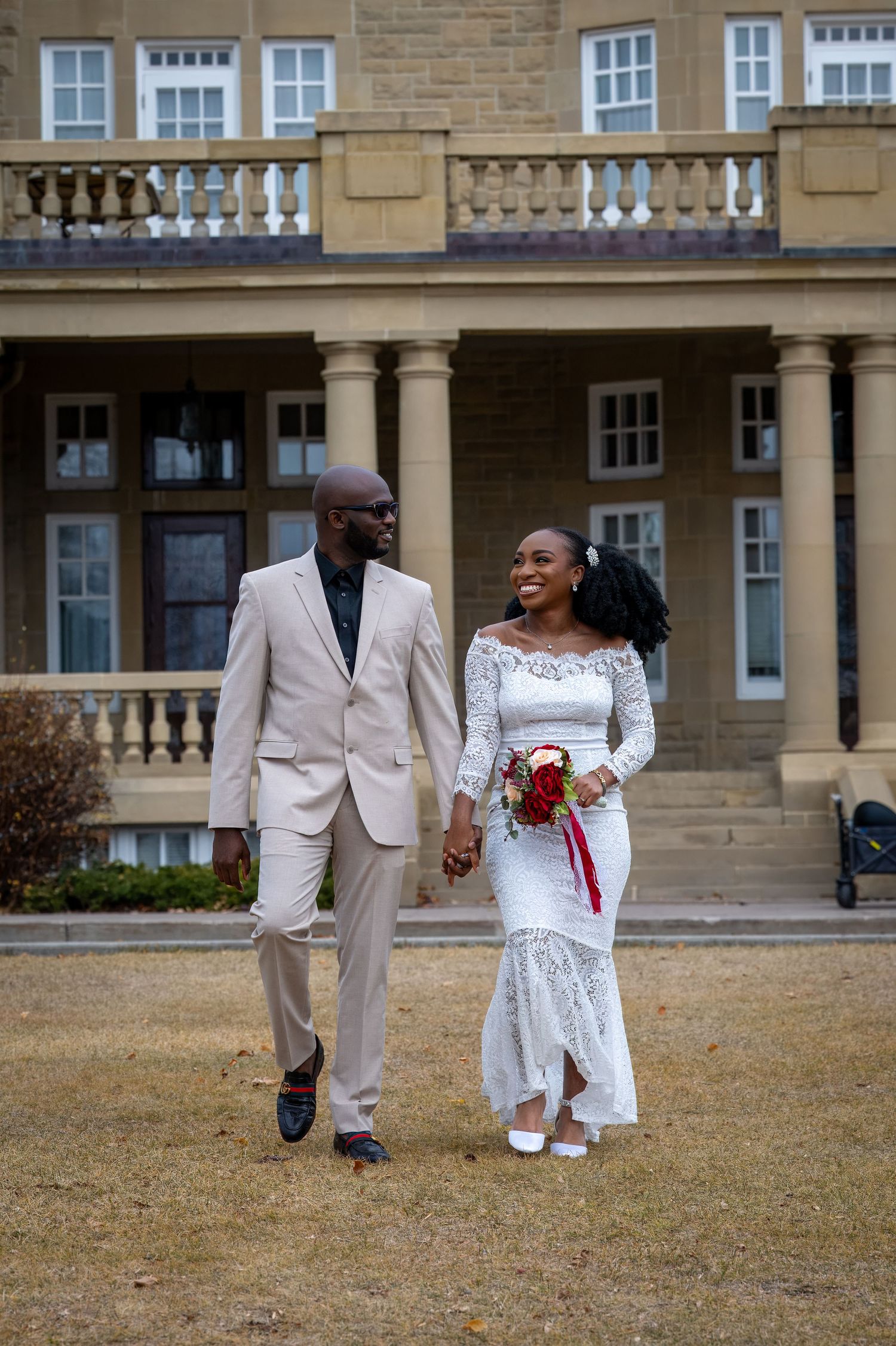 Wedding couple in white attire walking and posing on grounds of historic stone mansion.