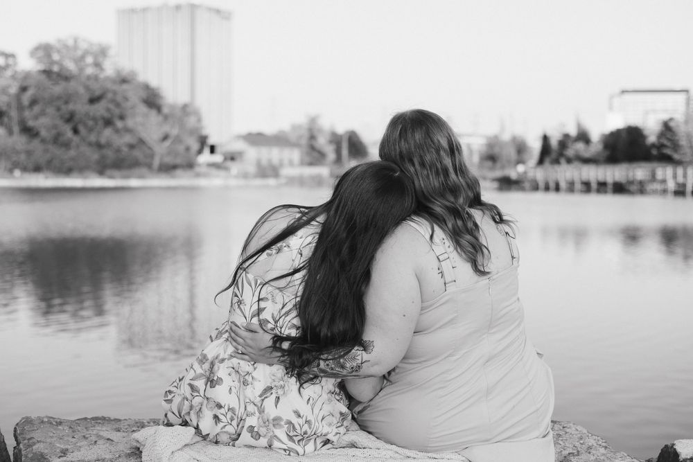 A person sits by a serene lake with long dark hair blowing in the wind against an urban backdrop in Waterloo Park