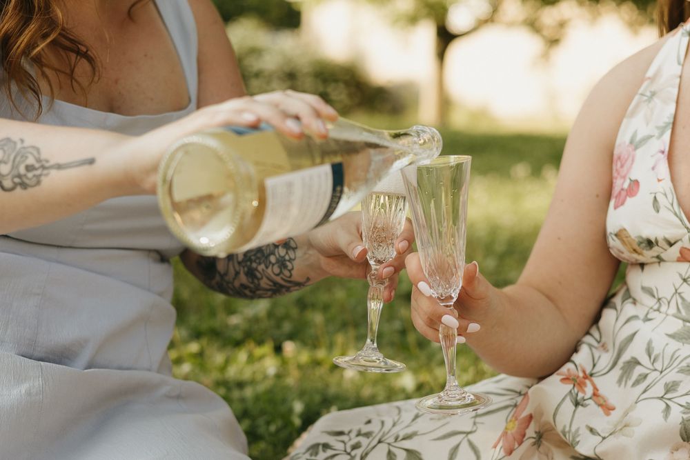 Two people pouring champagne into elegant flutes while sitting outdoors in a sunny garden setting.