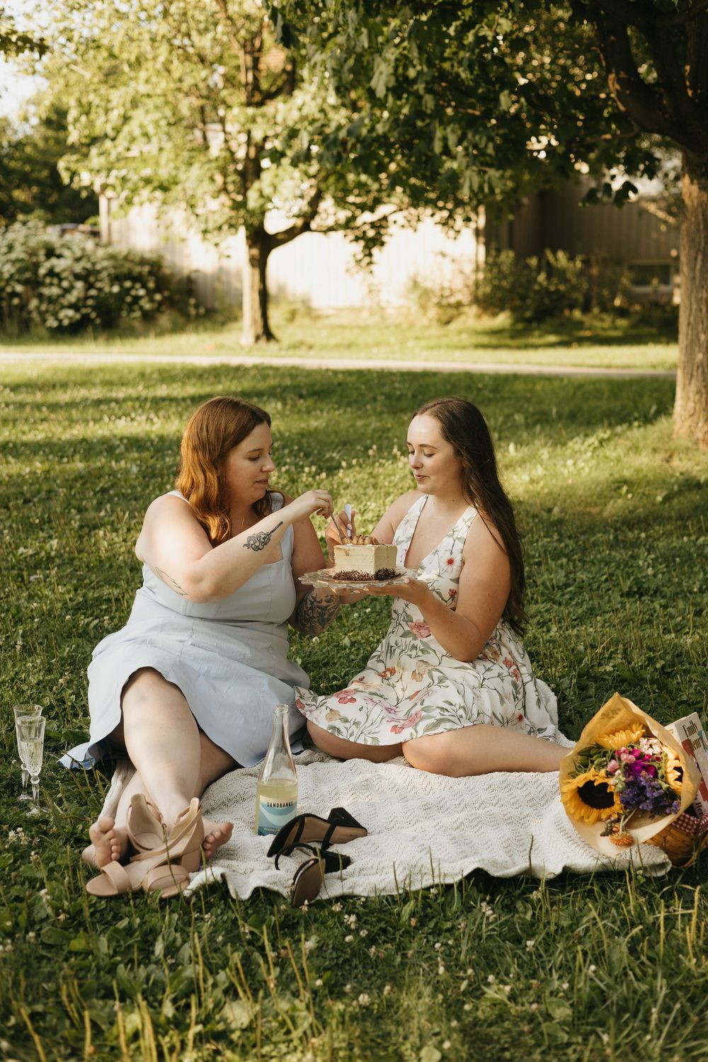 Two people in white dresses share an intimate picnic on grass with sunflowers and refreshments at golden hour.