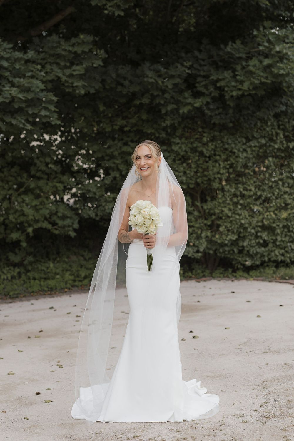 A bride in a flowing white wedding dress and veil holds a white bouquet while posing outdoors.