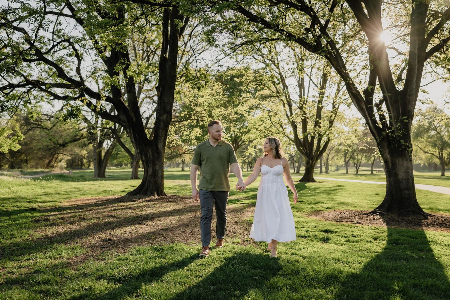 Couple holding hands walking through a sunlit park with large green trees during golden hour.