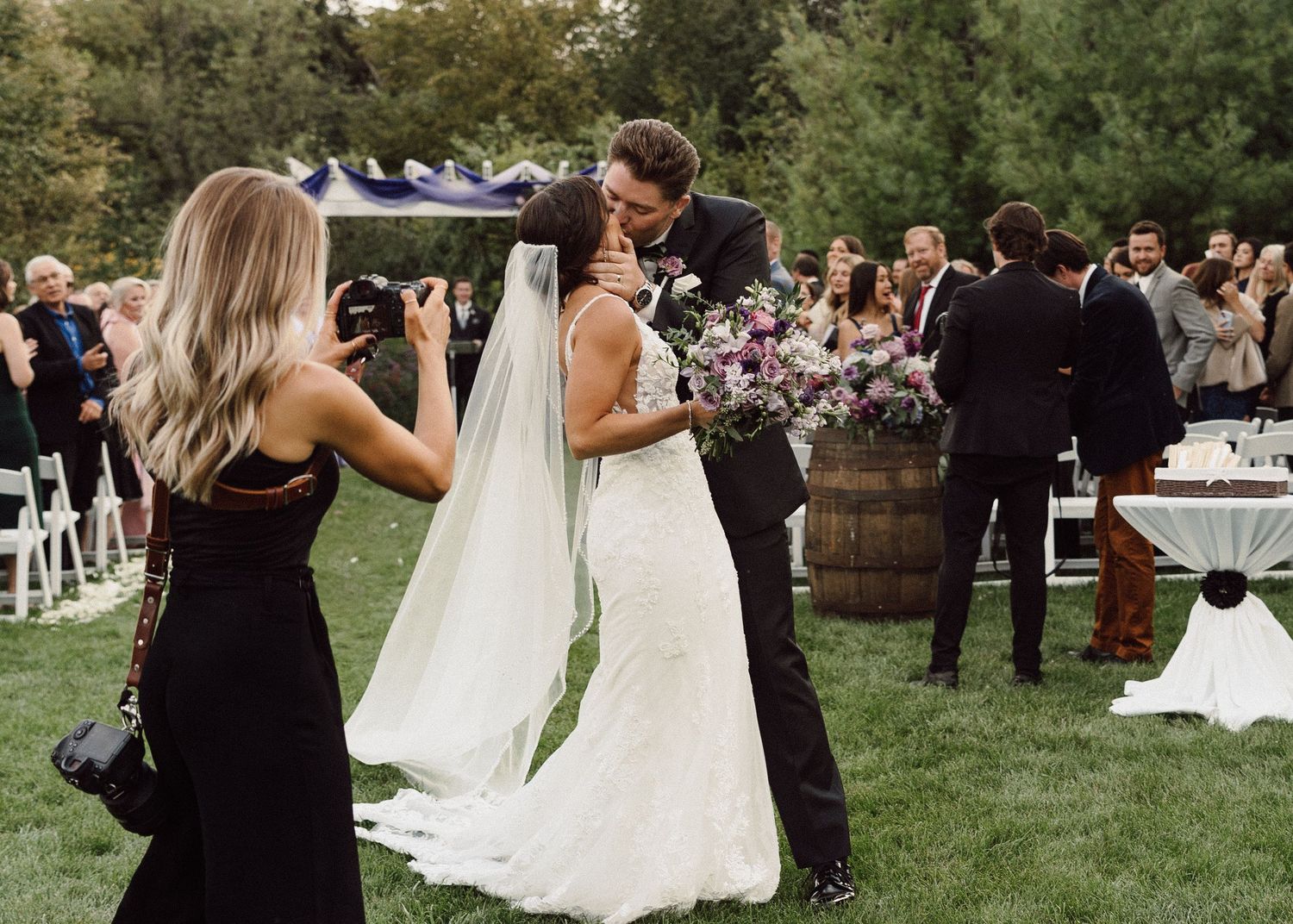 Outdoor wedding celebration with guests dancing on lawn near wine barrel and tables.