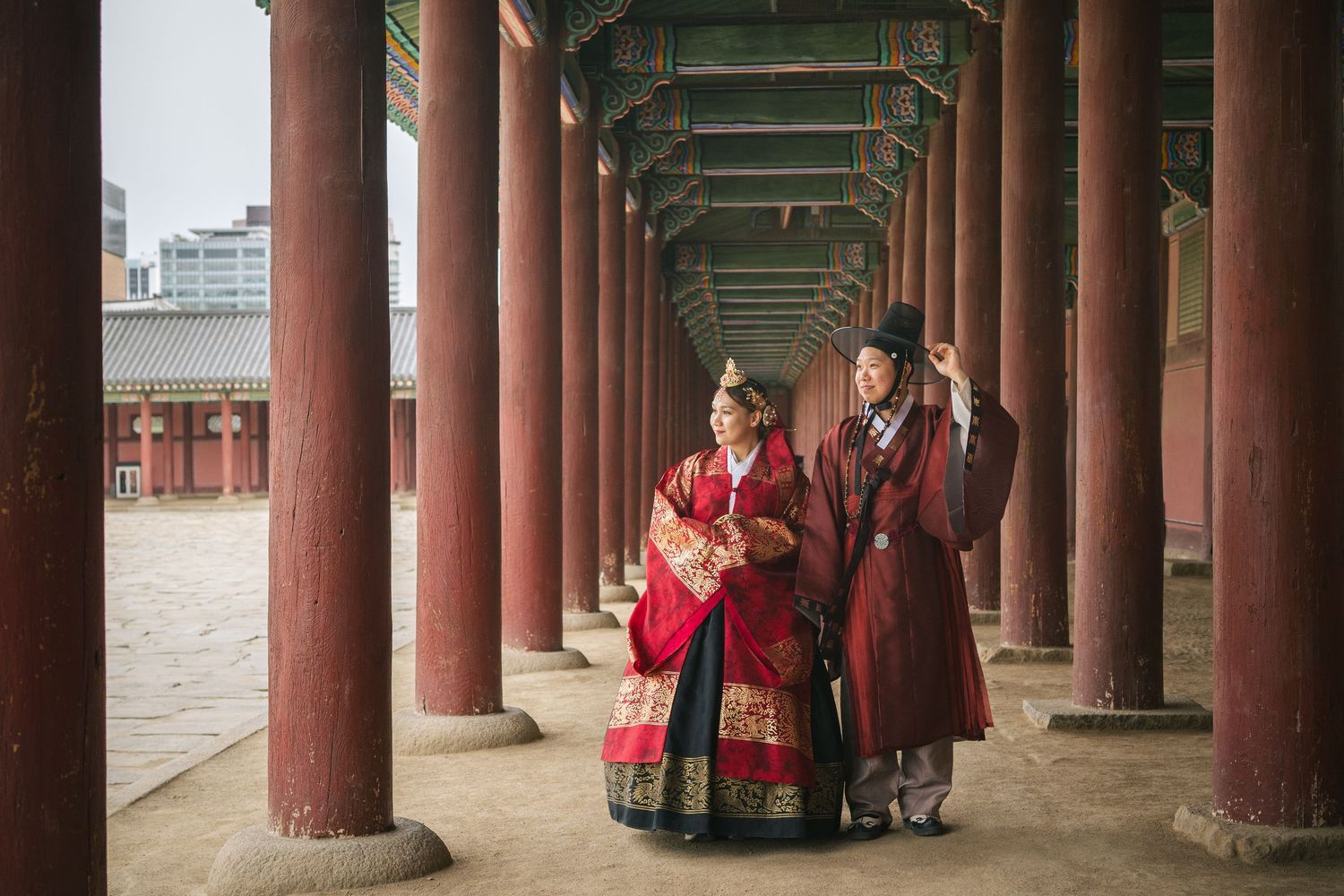 Traditional Korean palace courtyard with red wooden pillars and figures in traditional hanbok dress walking through corridor.