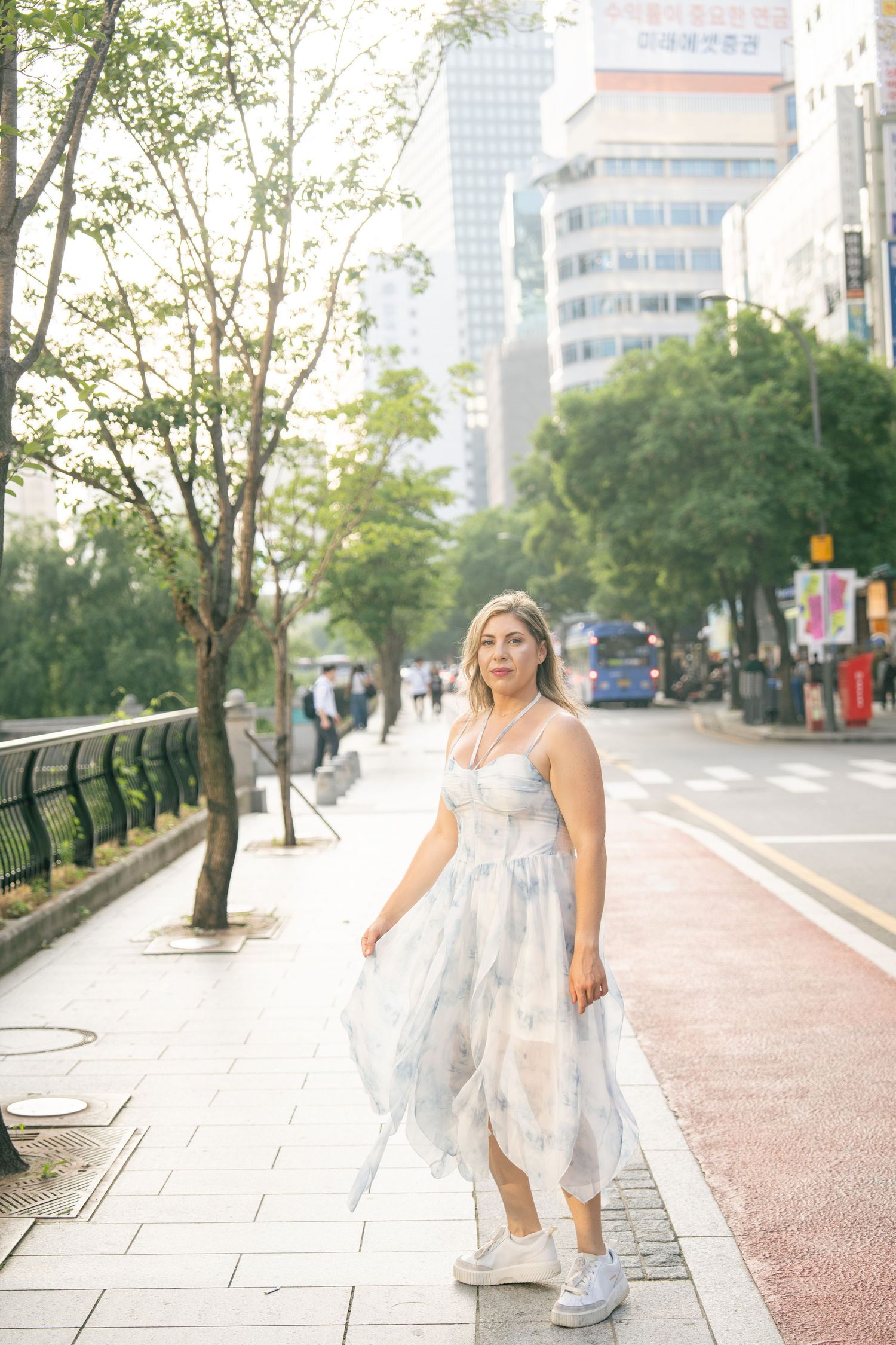A person in a flowy white dress poses gracefully along a tree-lined urban pathway with modern buildings in the background.