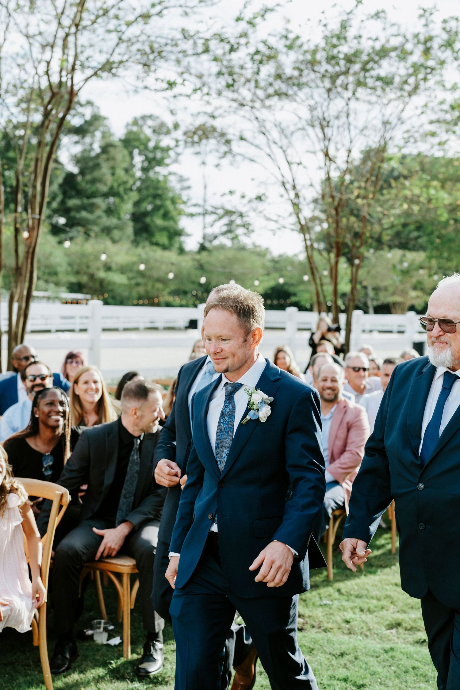 Wedding guests seated in wooden chairs on a grassy lawn during an outdoor ceremony.