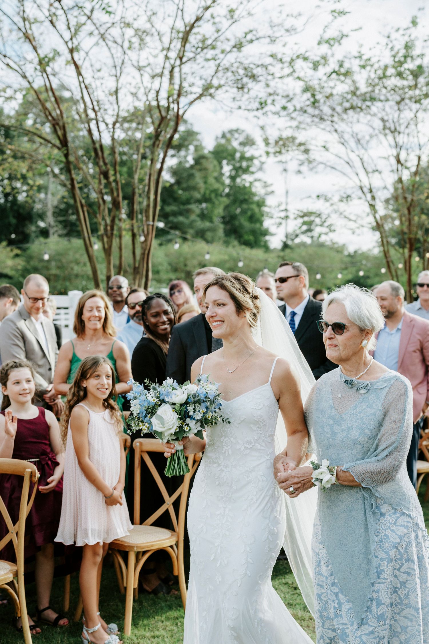 Elegant outdoor wedding ceremony with guests gathered in wooden chairs surrounded by lush greenery and trees.