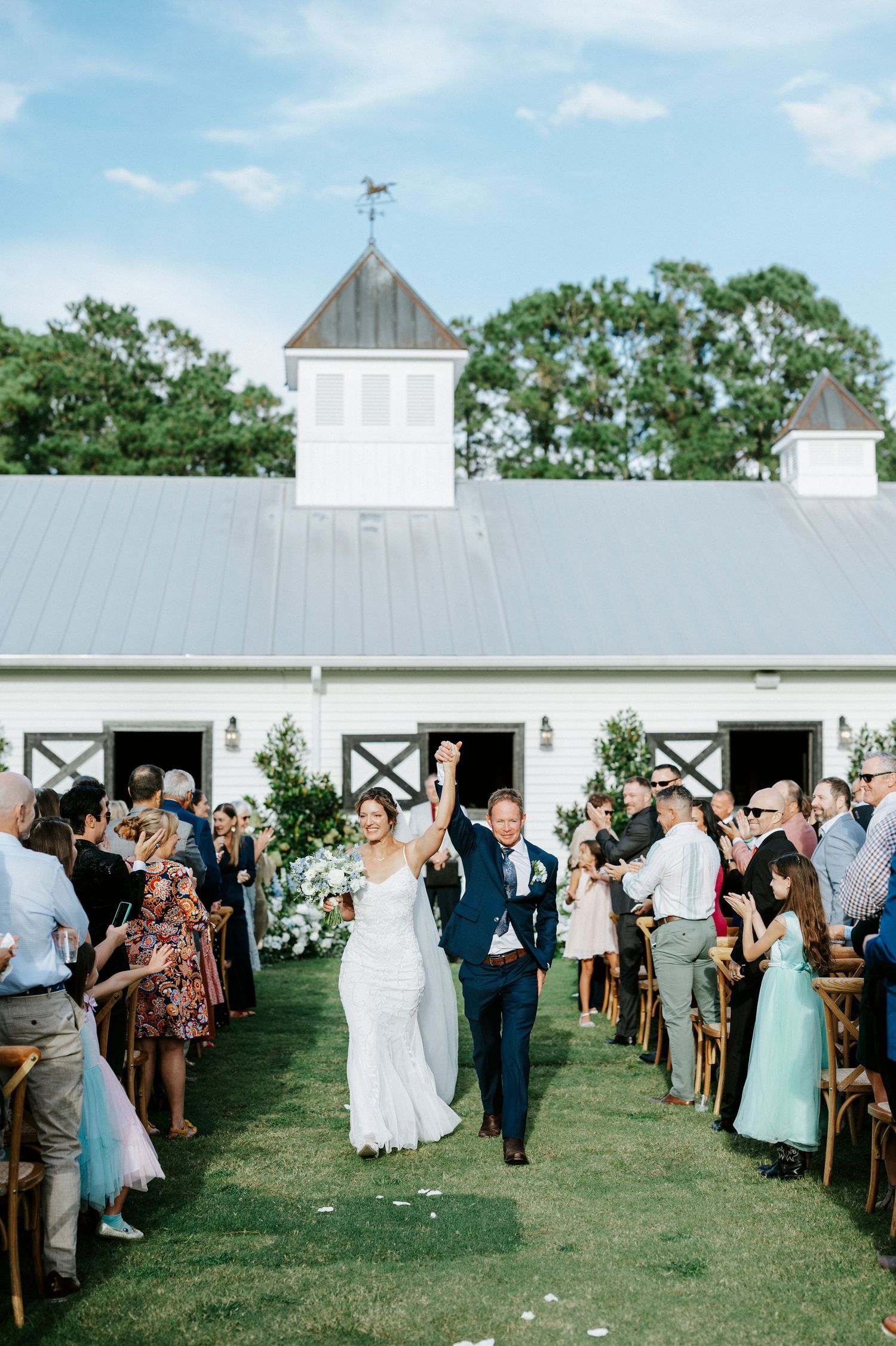 Wedding guests watch as a couple walks down the aisle at an outdoor barn venue with white buildings and green lawn.
