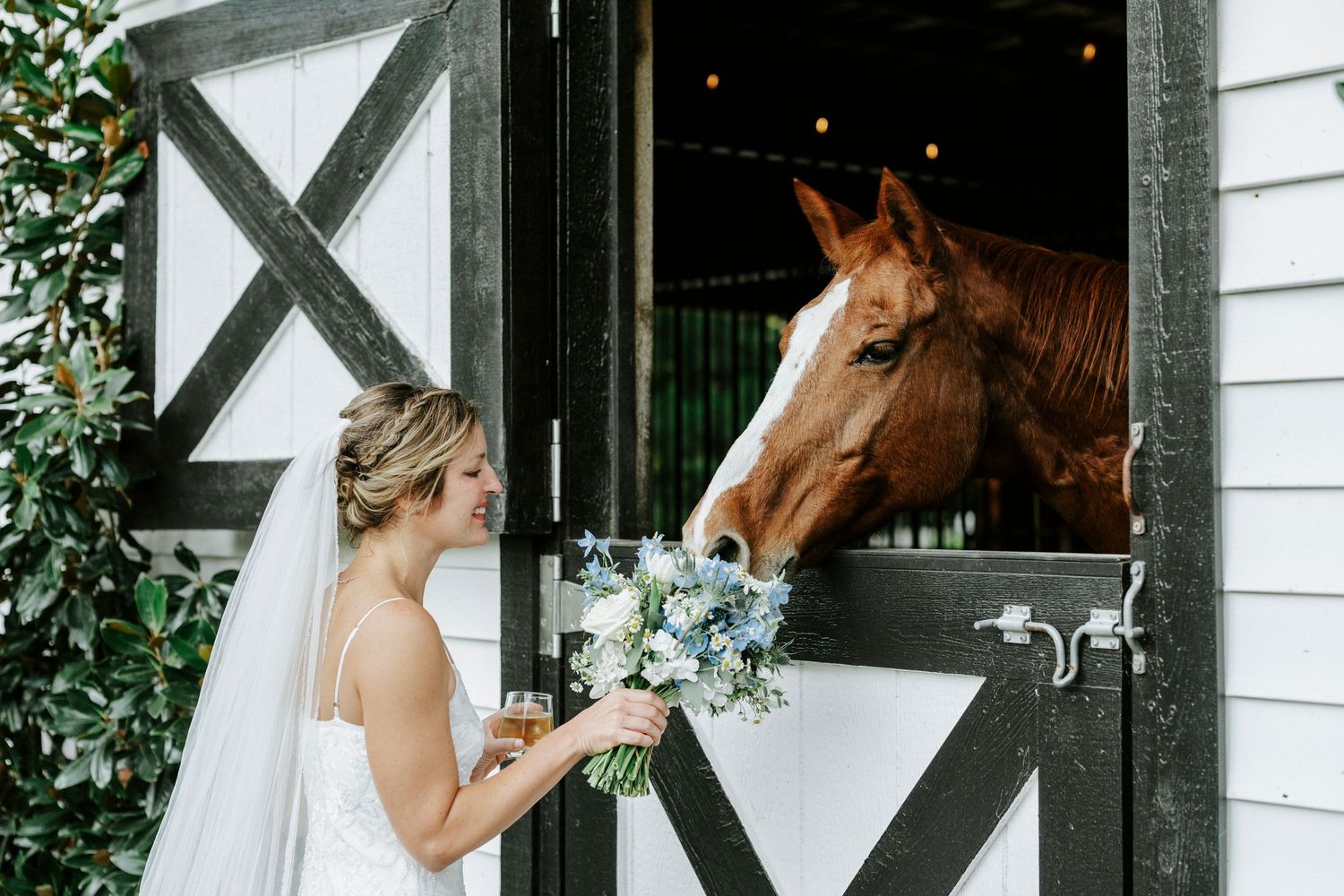 A bride in a white wedding dress holds her bouquet near a chestnut horse peeking out from a barn door.