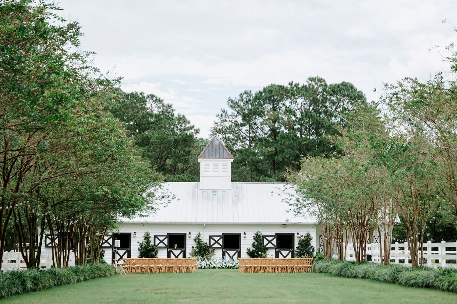 A panoramic view of a white church building with steeple surrounded by green trees and landscaping on a sunny day.