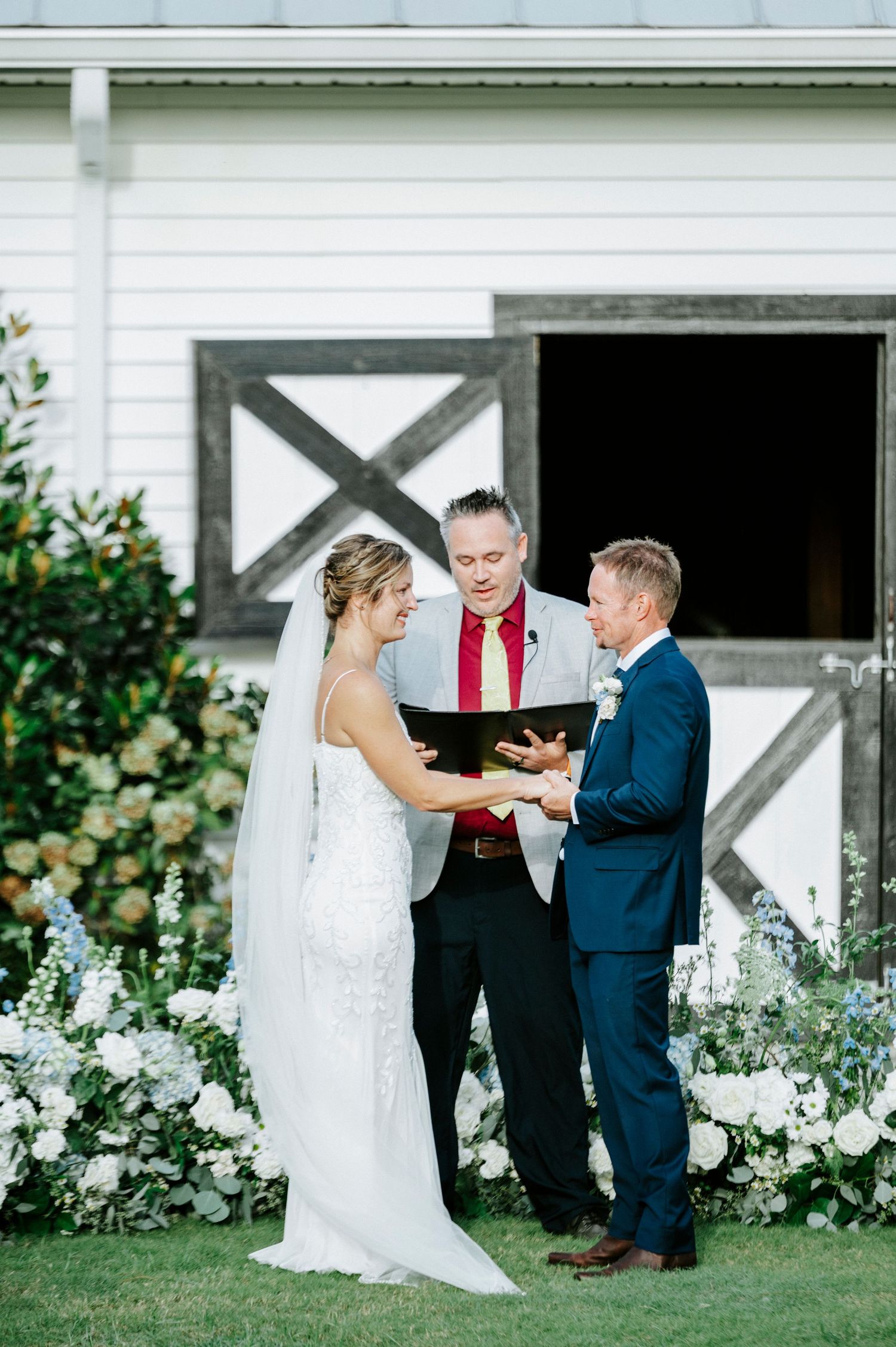 Wedding ceremony taking place in front of a white barn with floral decorations and greenery.