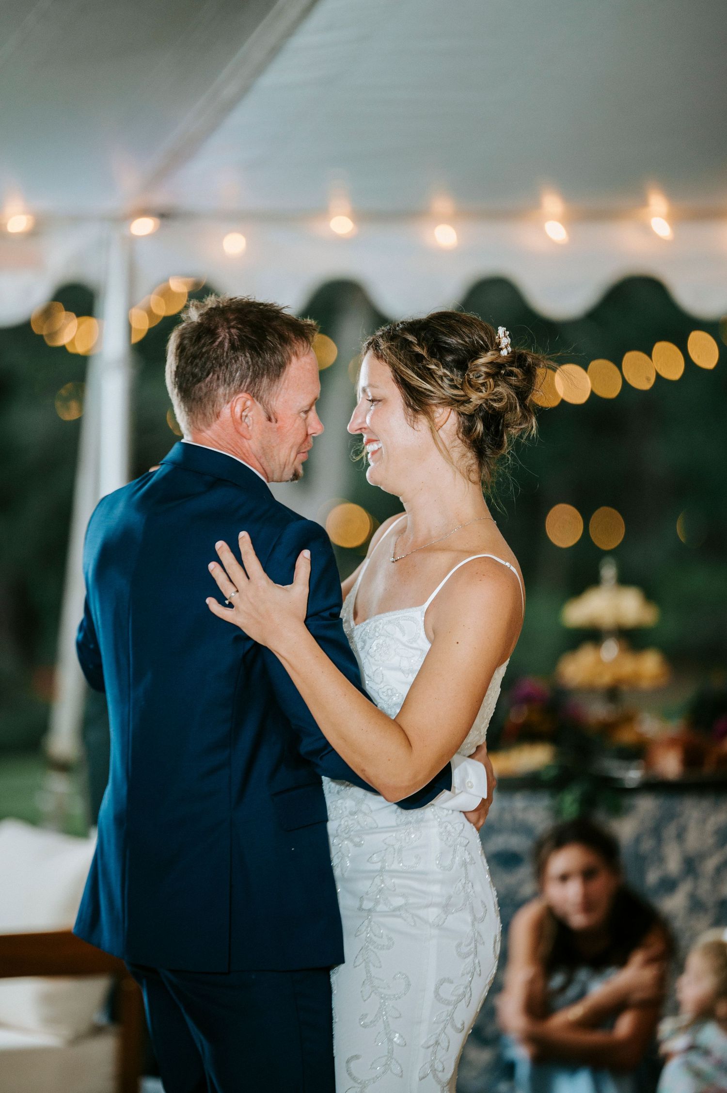 A couple shares their first dance as newlyweds at an indoor wedding reception with string lights in the background.