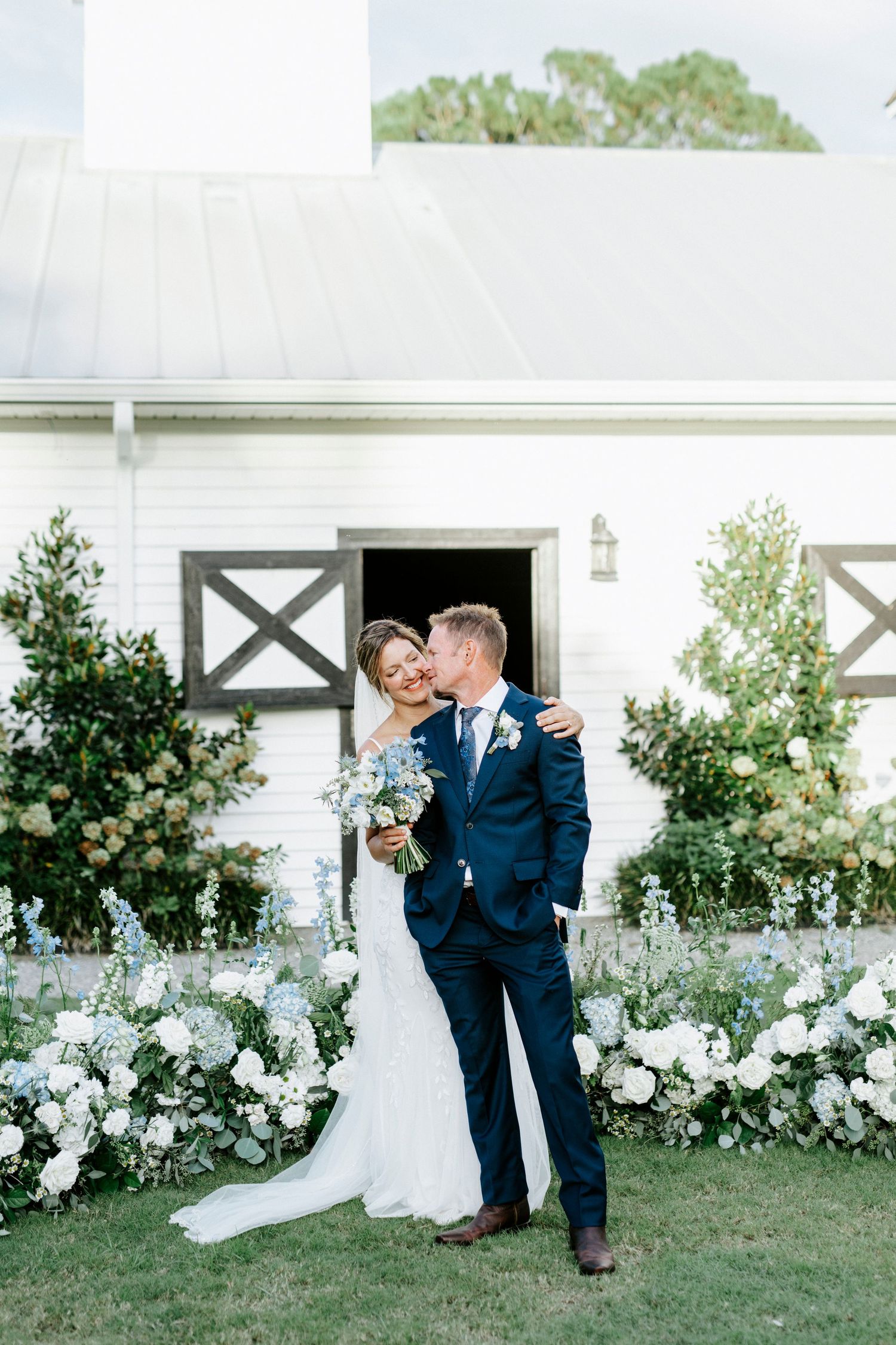 A couple shares a romantic moment in front of a white barn wall adorned with elegant floral arrangements.