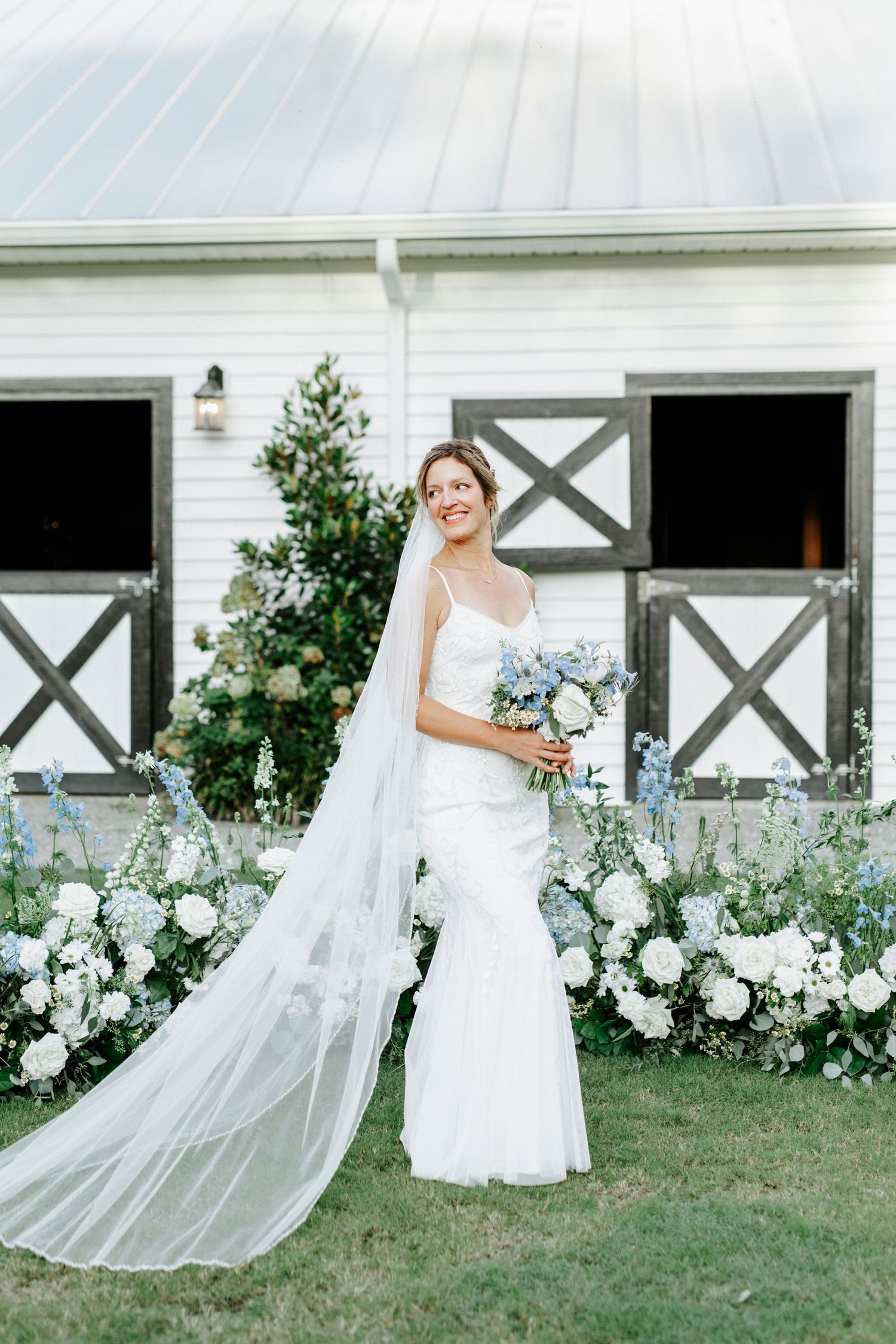 Elegant bride in flowing white wedding dress poses in front of rustic white barn with garden flowers.