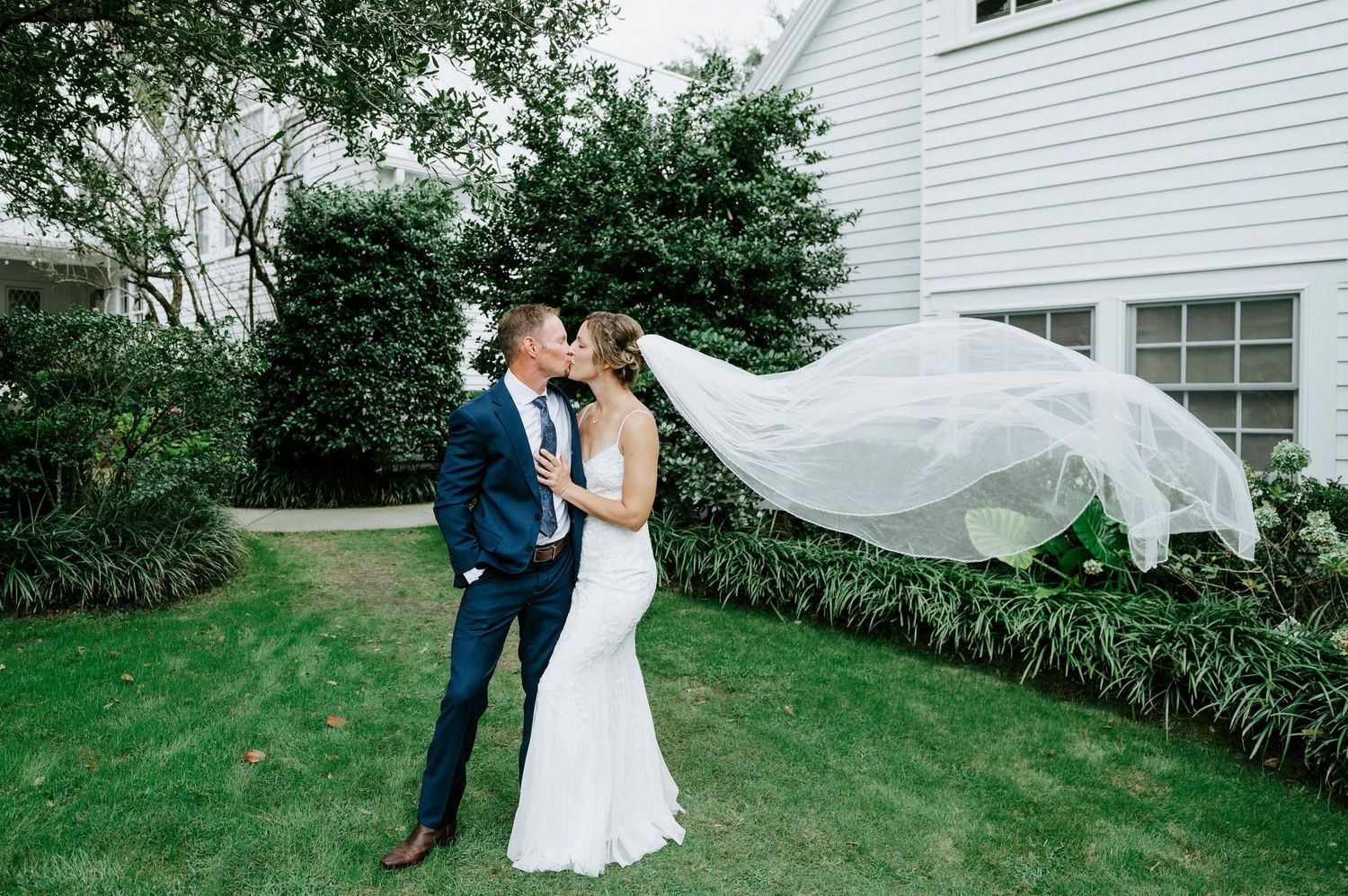 A couple in wedding attire stands together on a grassy lawn outside a white house during their outdoor celebration.