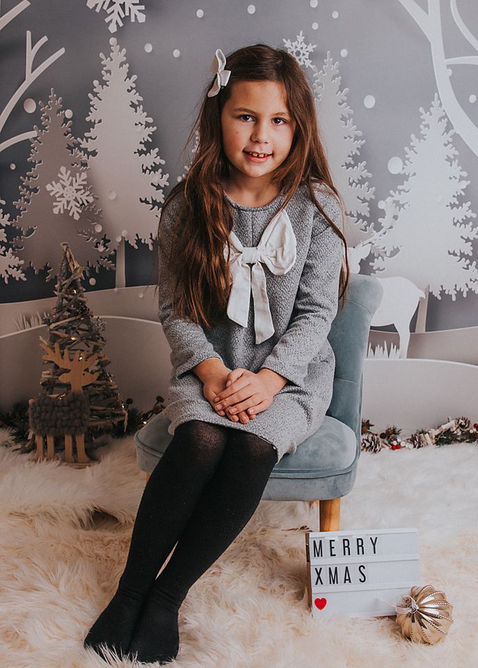 A child sits in a cozy winter wonderland setup with white trees, snowflakes, and a 'Merry' sign for holiday photos.