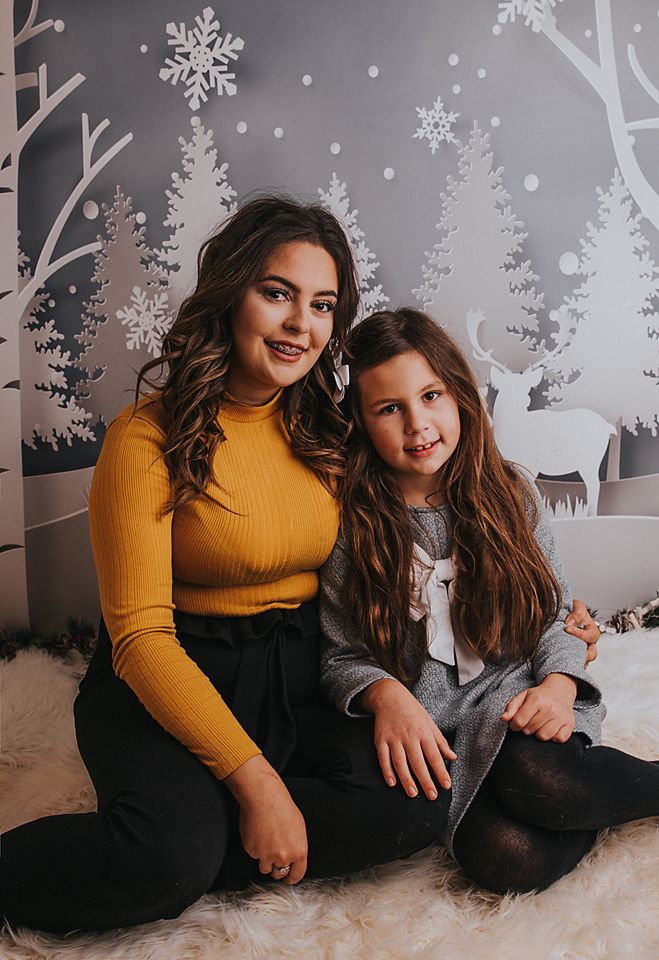 Two people in winter outfits sit together and laugh against a white wall decorated with snowflake and tree decals.