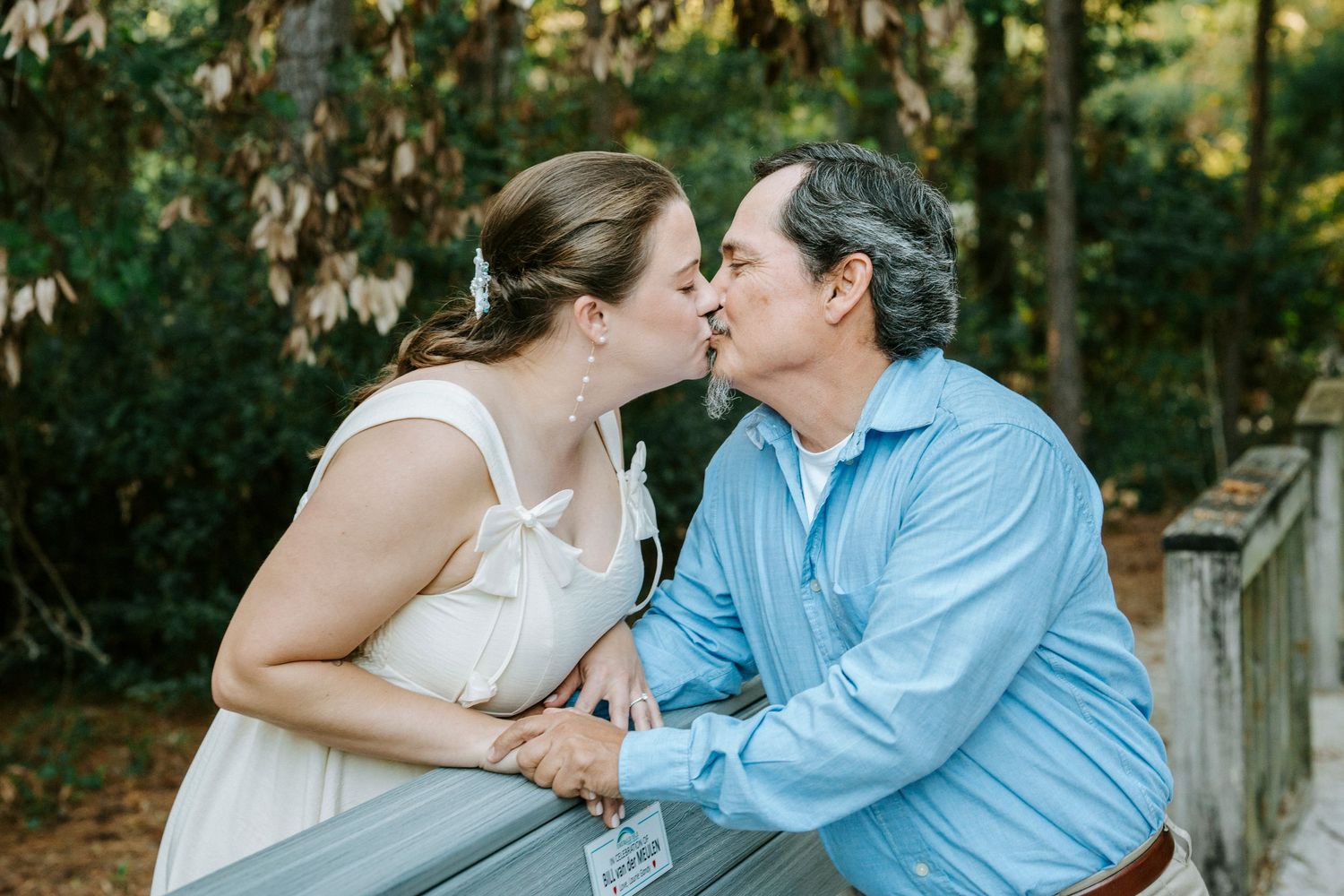 A romantic couple in white and blue attire share tender kisses while sitting outdoors in a garden setting.