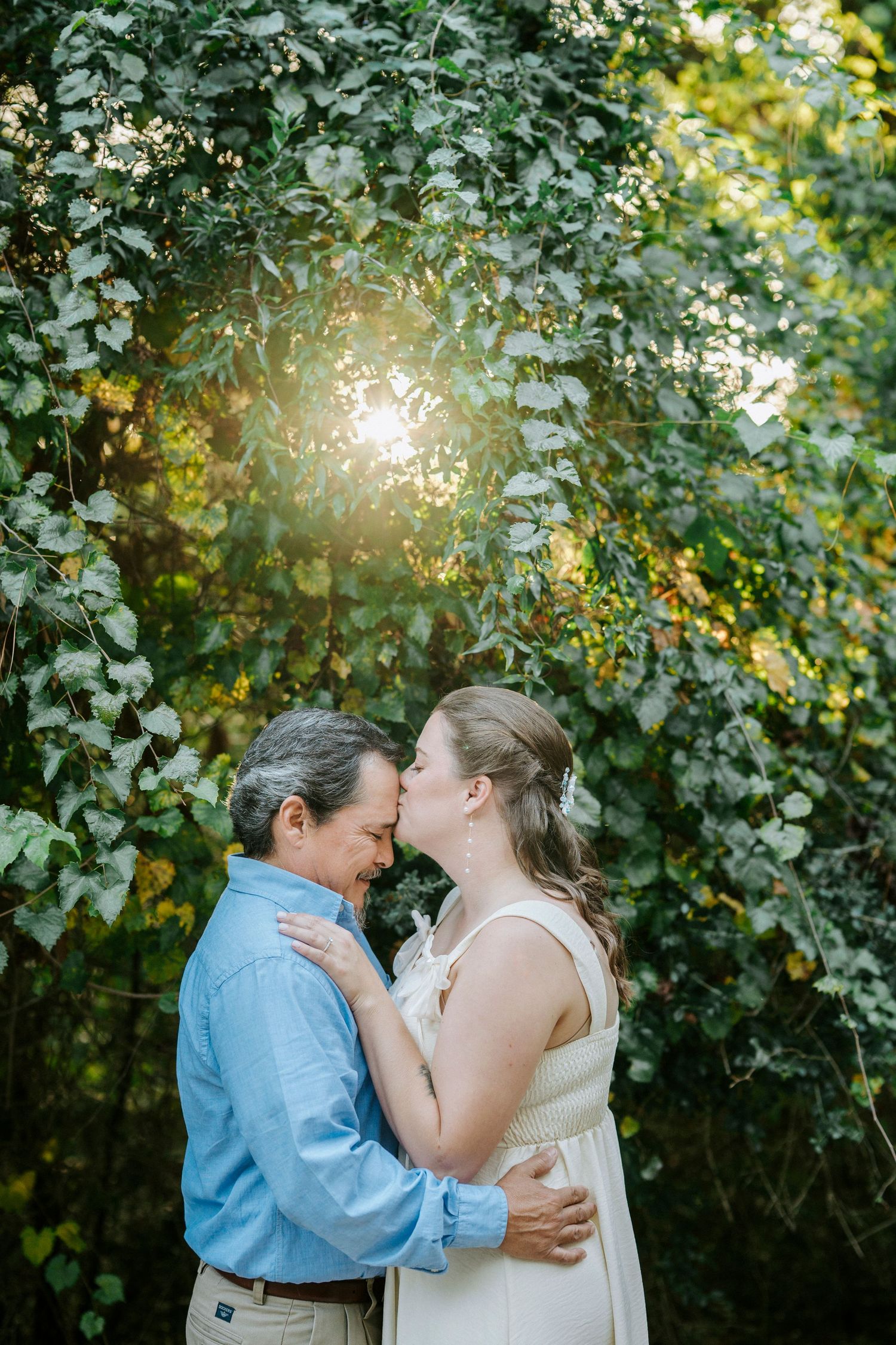 A couple embraces in a sunlit garden with lush greenery and golden sunlight filtering through.