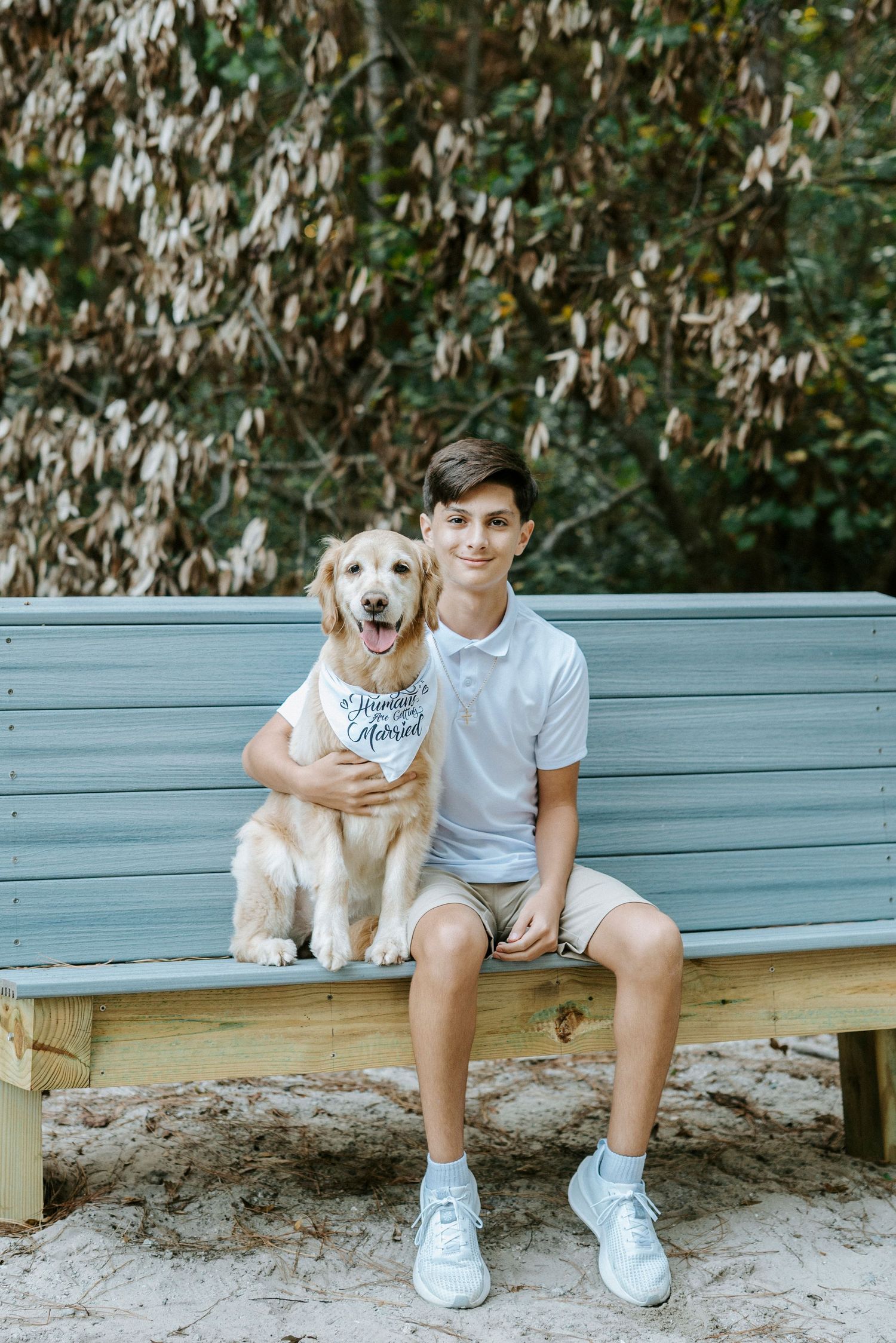 Golden retriever wearing a bandana sits with companion on a blue wooden bench in an outdoor setting.