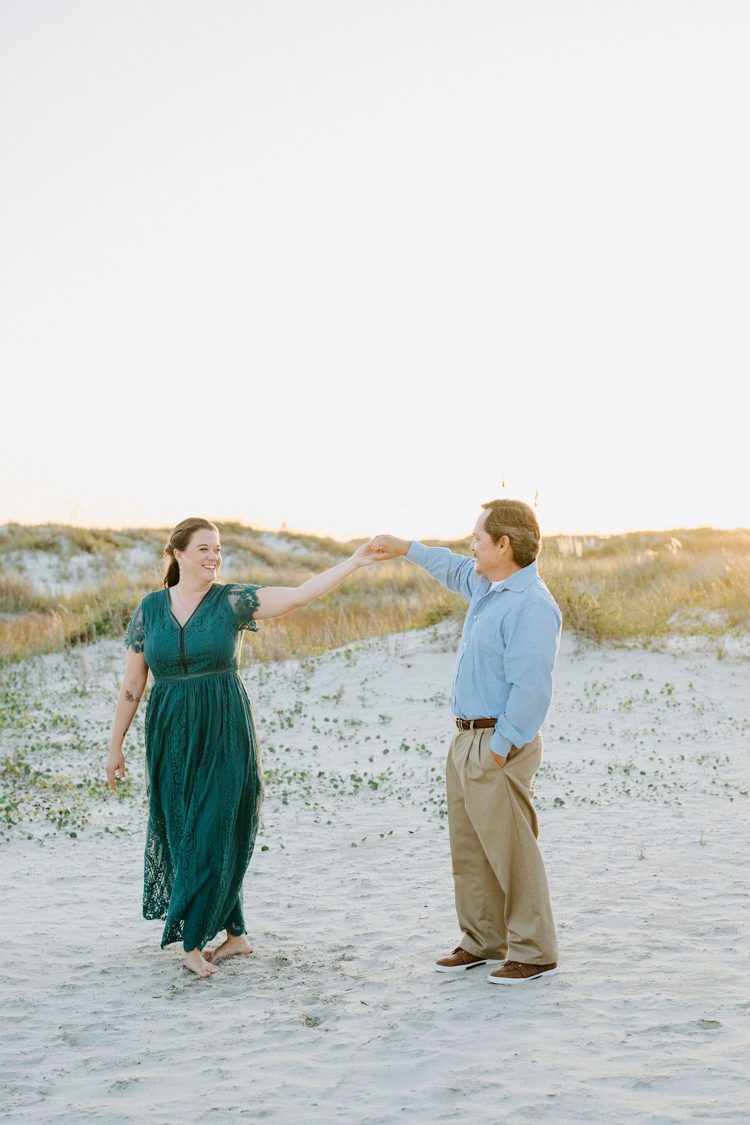 A couple dances playfully on a sandy beach at sunset wearing blue and green outfits.